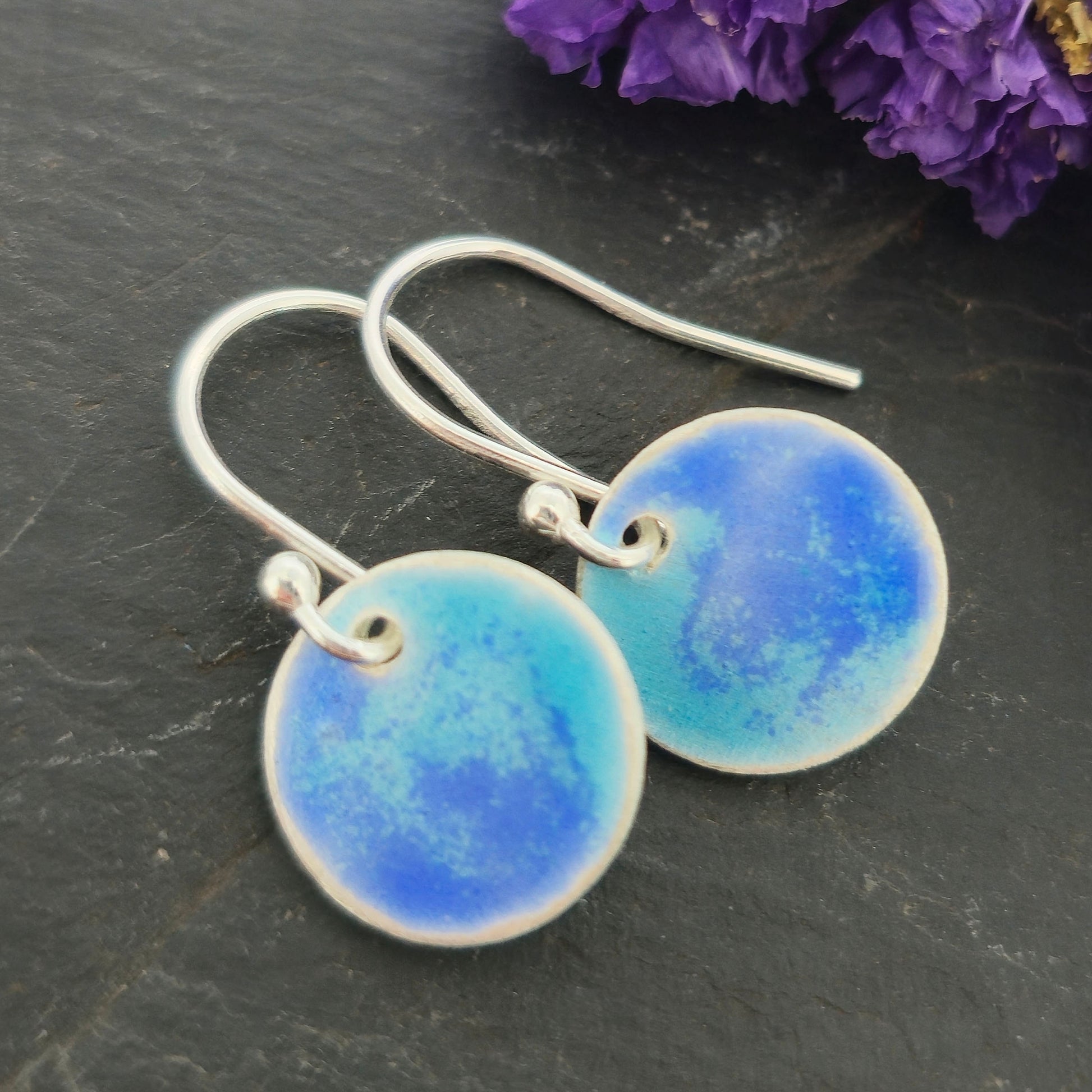 Handmade silver round drop earrings with a mix of dark and light blue enamel. Pictured on a black background with purple flowers.