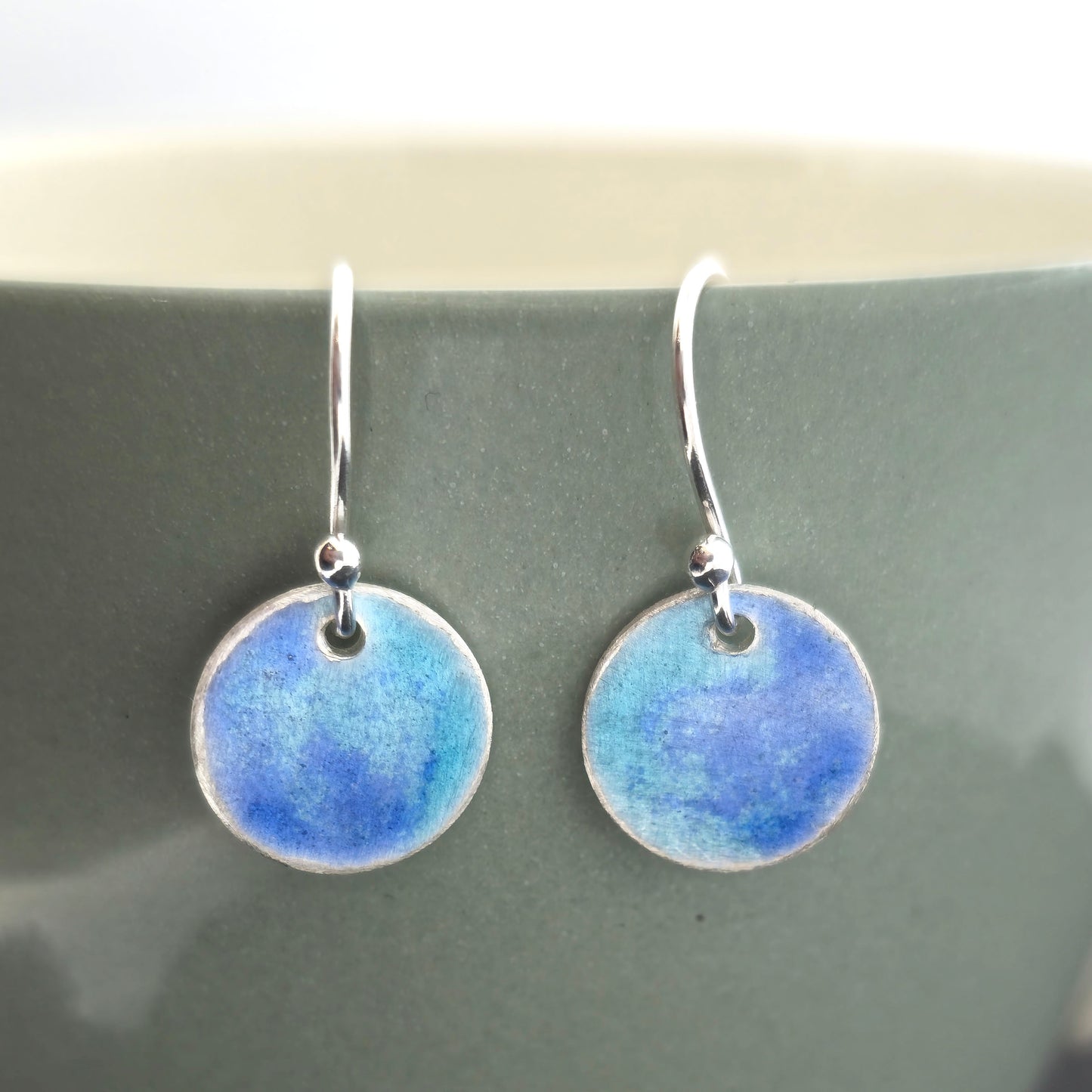 Handmade silver round drop earrings with a mix of dark and light blue enamel. Pictured on a pale green cup. Mijoux Jewellery.