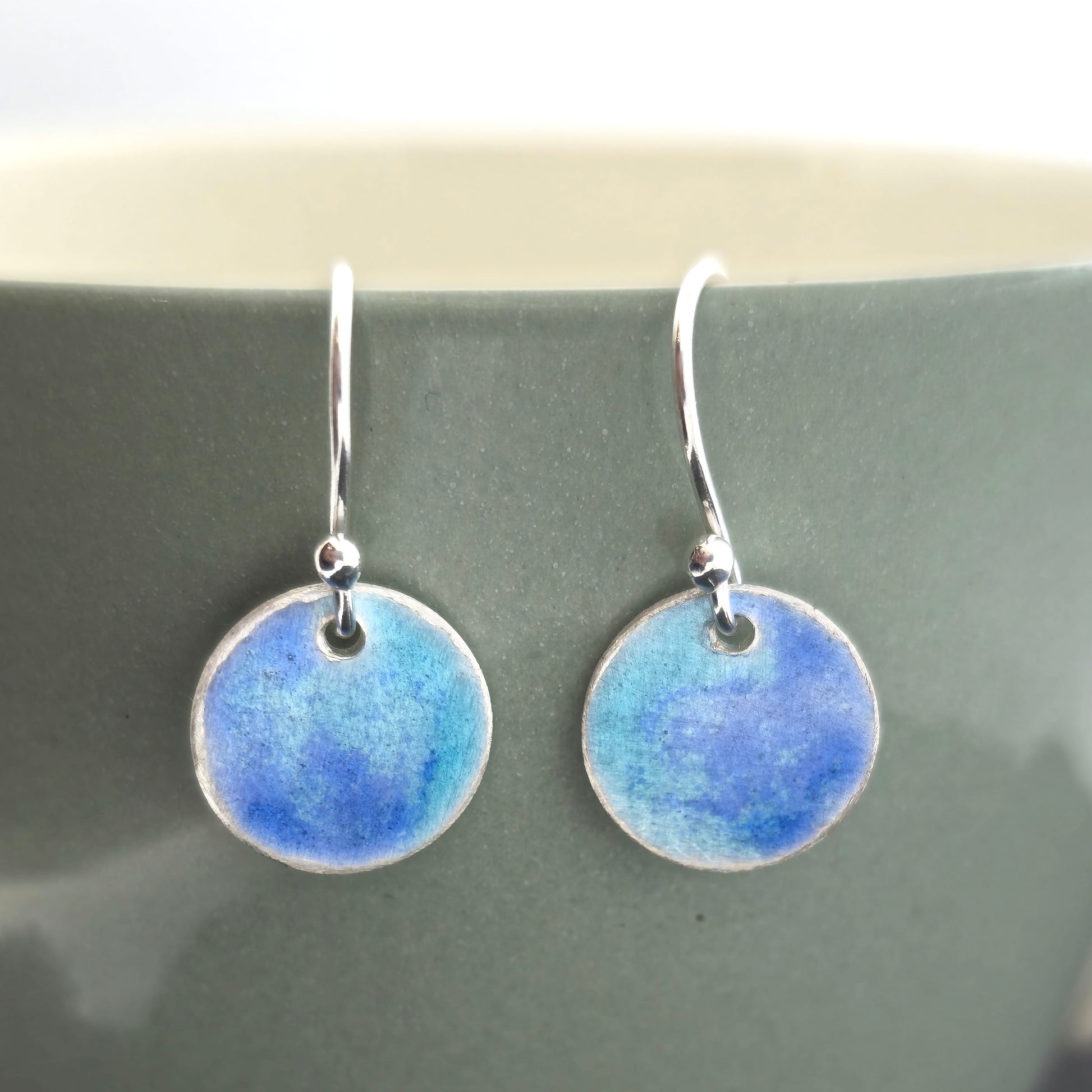 Handmade silver round drop earrings with a mix of dark and light blue enamel. Pictured on a pale green cup. Mijoux Jewellery.