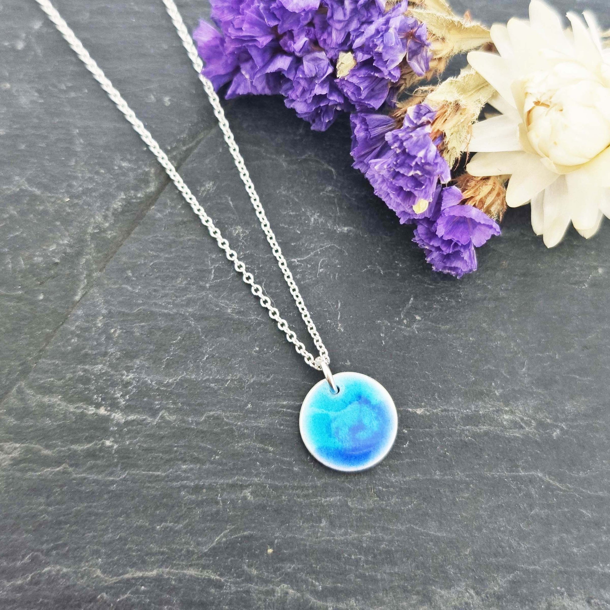 A round silver pendant with a mix of light and dark blue enamel suspended from a silver chain. With flowers.