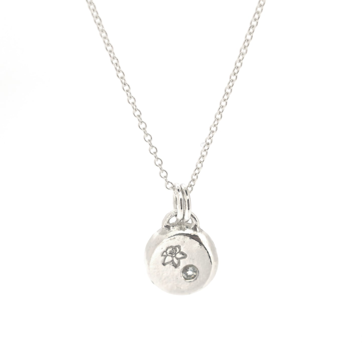 A round silver flat pebble pendant with a daffodil engraved on it and a flush set pale blue aquamarine gemstone.