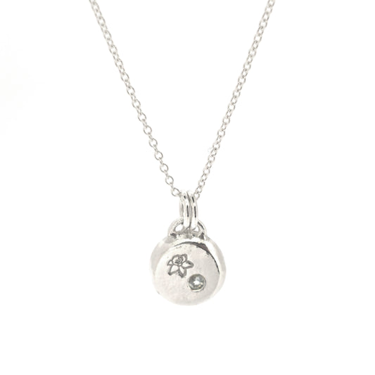 A round silver flat pebble pendant with a daffodil engraved on it and a flush set pale blue aquamarine gemstone.