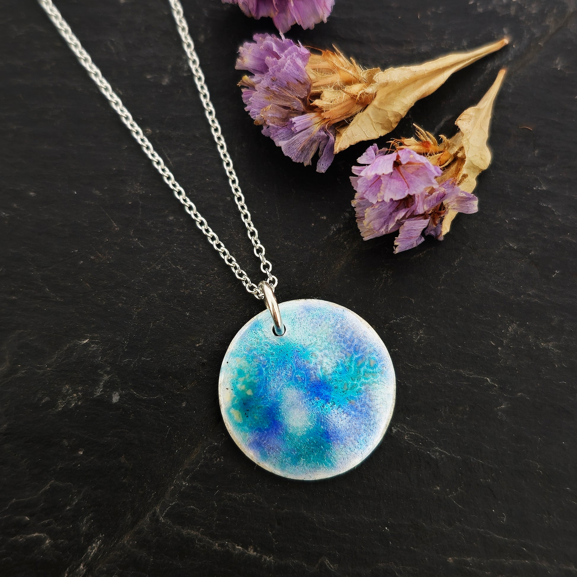 A round silver pendant on a silver chain. The pendant is covered in turquoise, blue and clear enamel. Shown on slate with flowers.