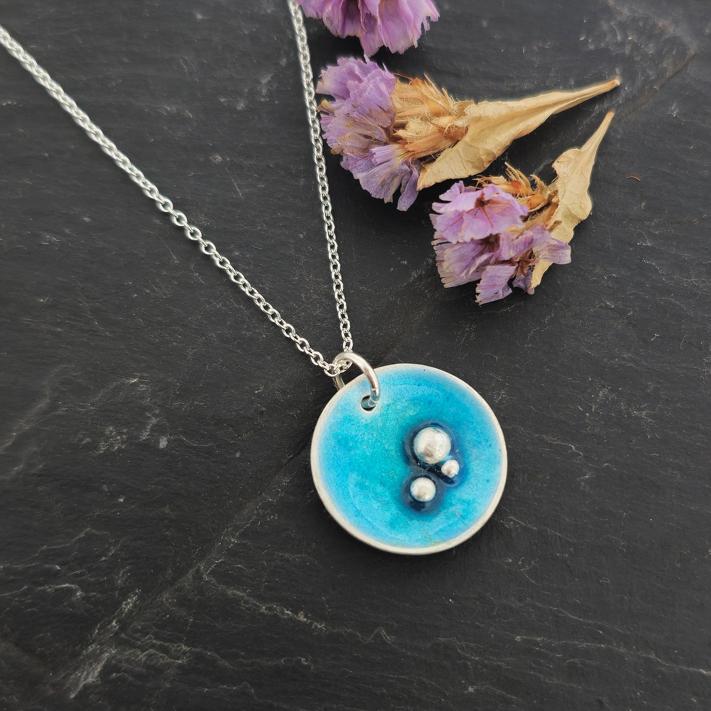 A round silver pendant on a silver chain. The pendant has 3 silver balls off-centre and is covered in turquoise blue enamel. Shown on slate with flowers.