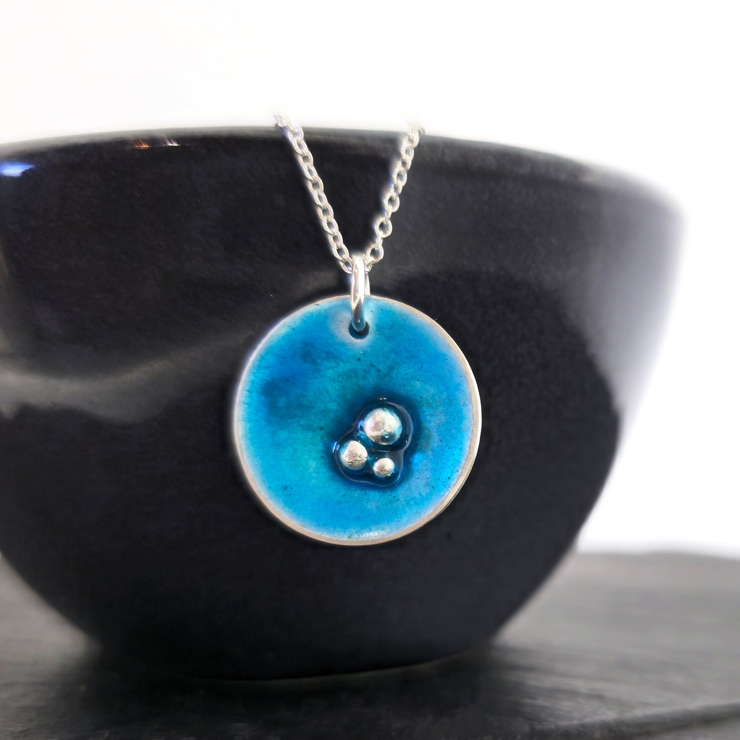 A round silver pendant on a silver chain. The pendant has 3 silver balls off-centre and is covered in turquoise blue enamel. Shown on a black bowl.