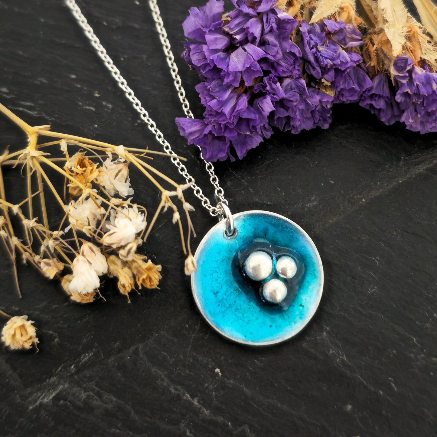A handmade round silver pendant on a silver chain. The pendant has 3 silver balls off-centre and is covered in turquoise blue enamel. Mijoux Jewellery.