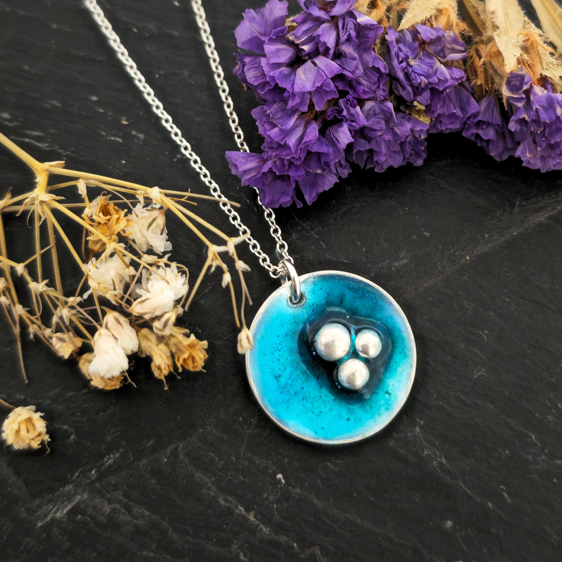 A handmade round silver pendant on a silver chain. The pendant has 3 silver balls off-centre and is covered in turquoise blue enamel. Mijoux Jewellery.