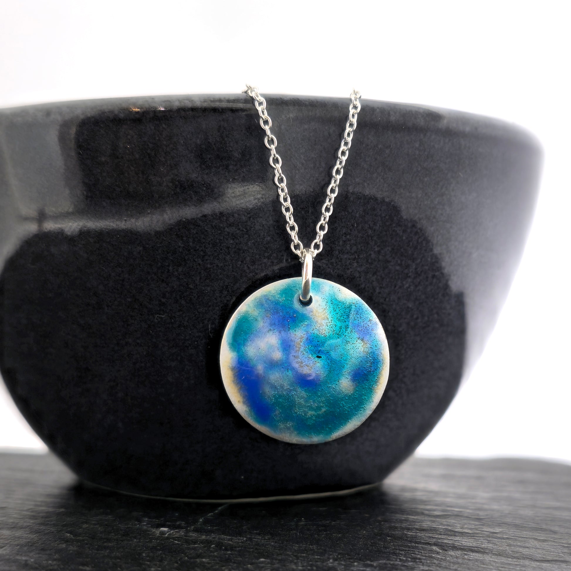 A round silver pendant on a silver chain. The pendant is covered in turquoise, blue and clear enamel. Shown on a black bowl.