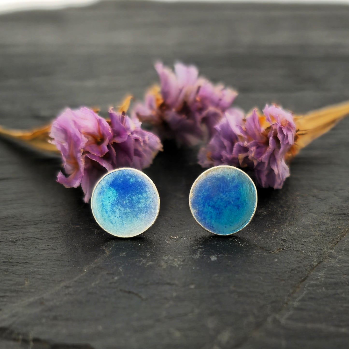 Round silver stud earrings with a mix of blue enamels. Shown on slate with flowers.