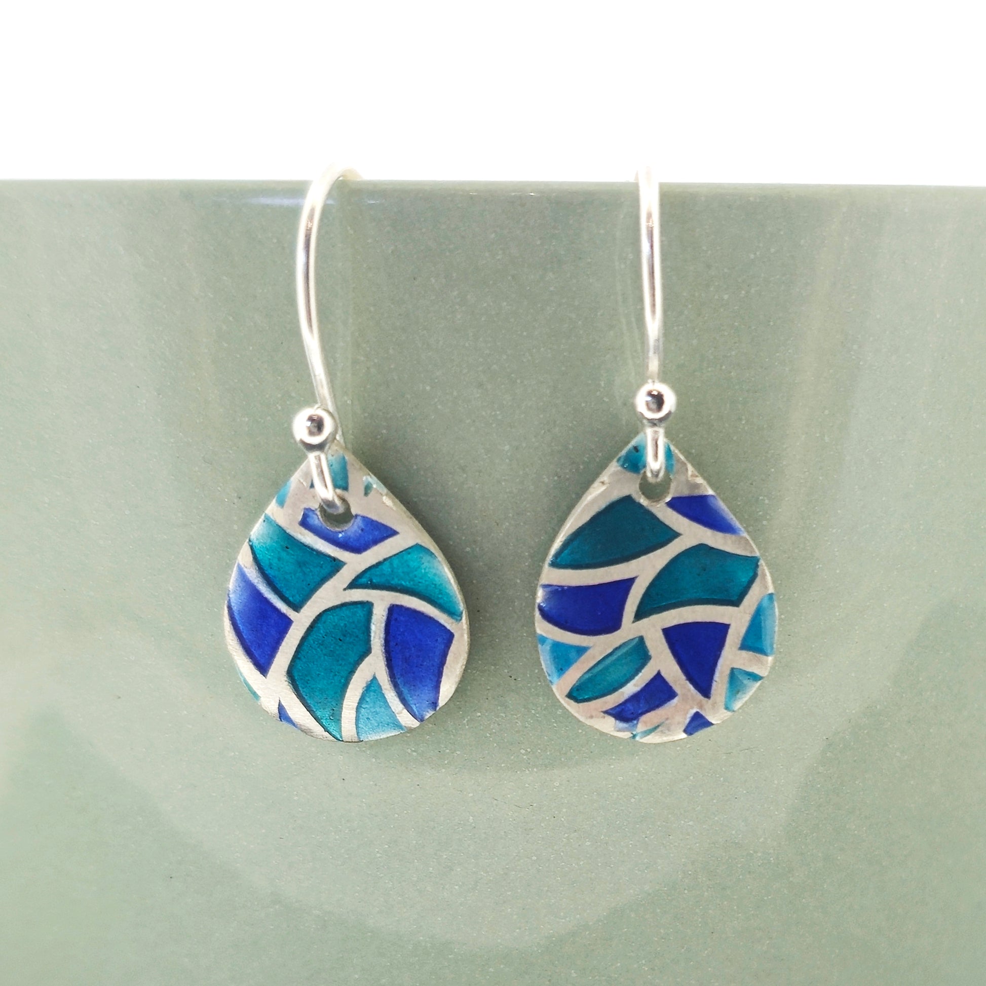 Teardrop shaped drop earrings featuring geometric cells of blue, turquoise and green enamel with silver outlines. Suspended on silver ear hooks. 