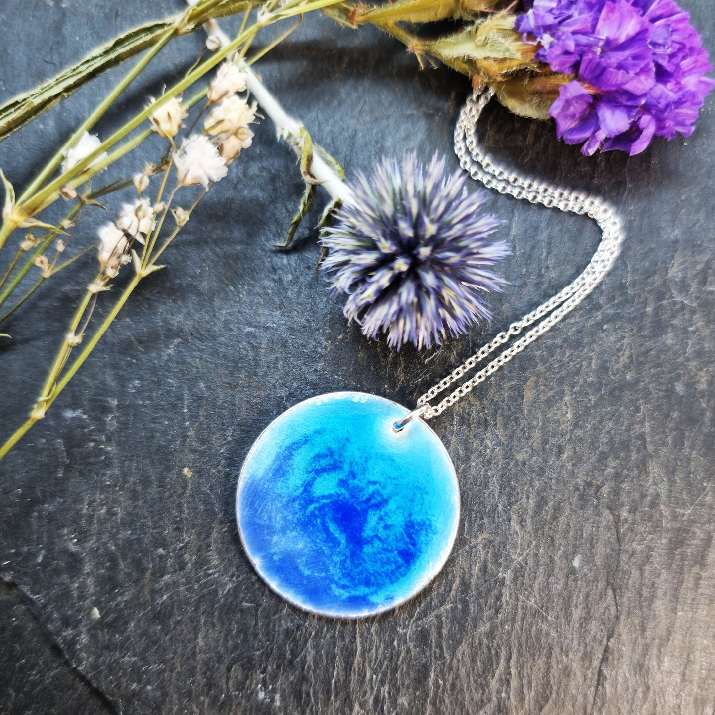 A round silver pendant with a mix of light and dark blue enamel suspended from a silver chain. With flowers