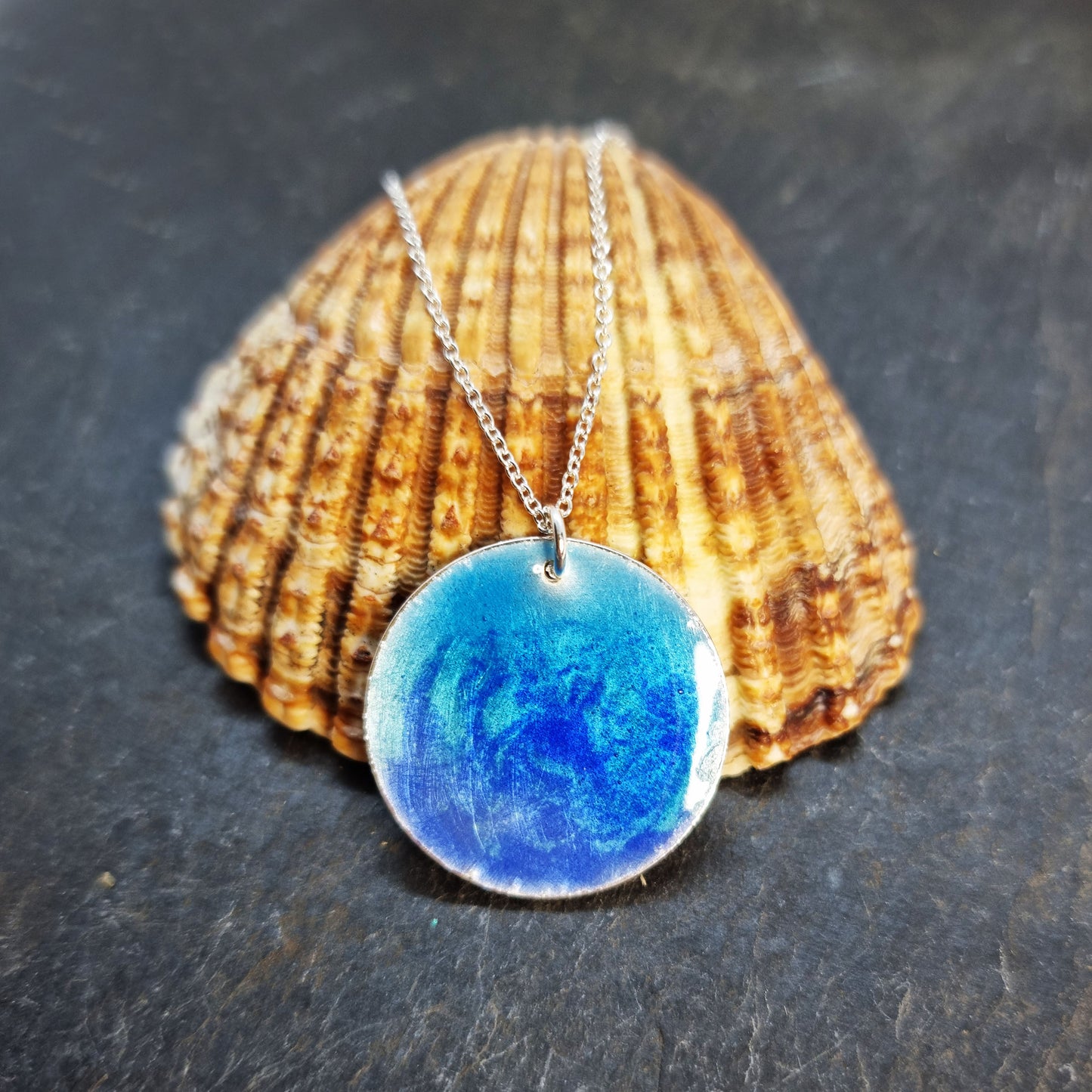 A round silver pendant with a mix of light and dark blue enamel suspended from a silver chain. On shell.