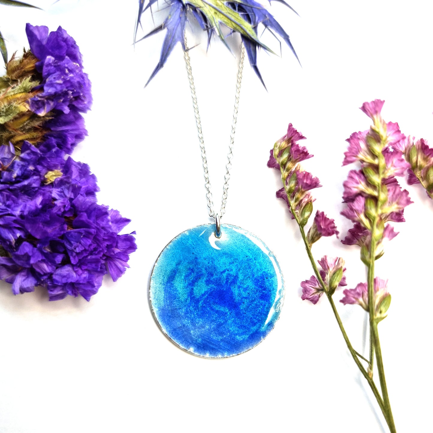 A round silver pendant with a mix of light and dark blue enamel suspended from a silver chain. With flowers.