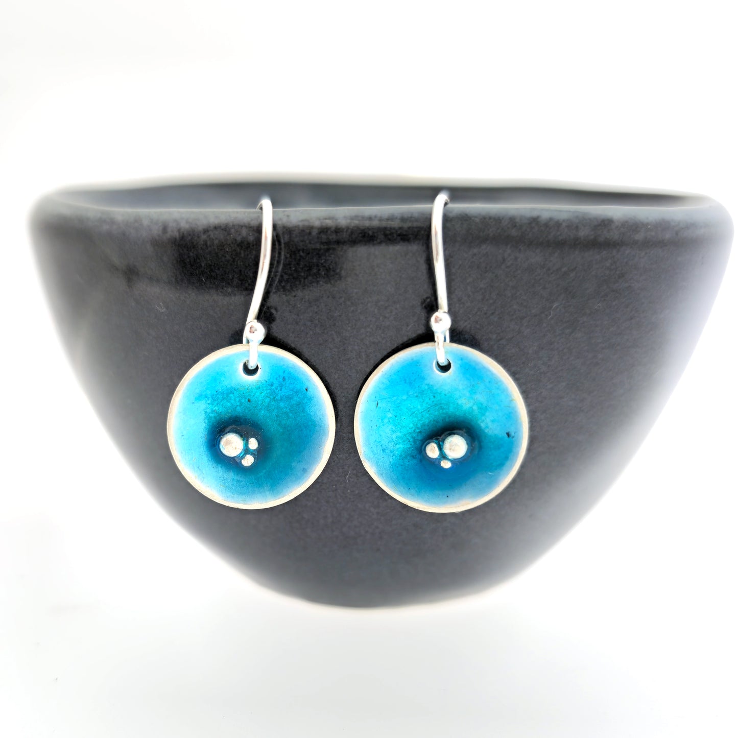 Round silver drop earrings. Each earring has 3 silver balls off-centre and a turquoise blue enamel. Suspended from silver ear hooks. Shown on a black bowl.