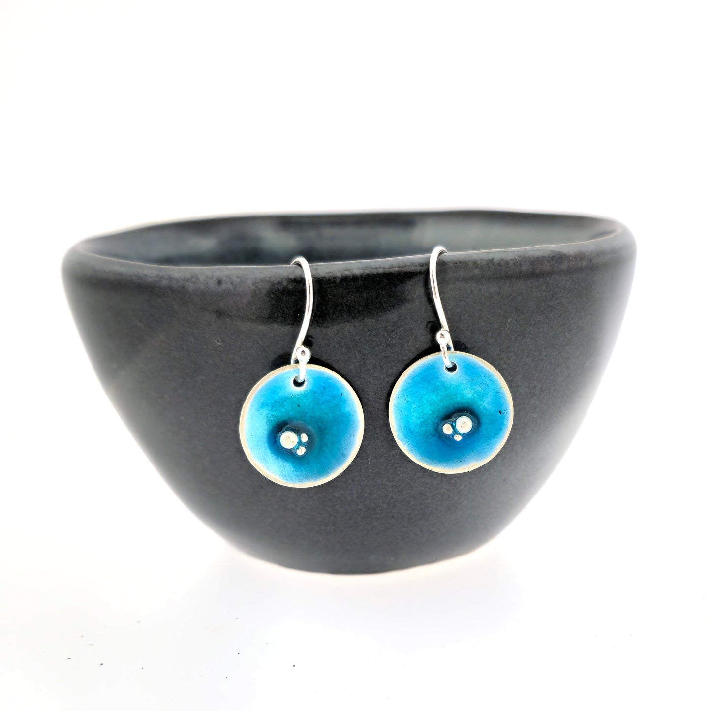 Round silver drop earrings. Each earring has 3 silver balls off-centre and a turquoise blue enamel. Suspended from silver ear hooks. Shown on a black bowl.