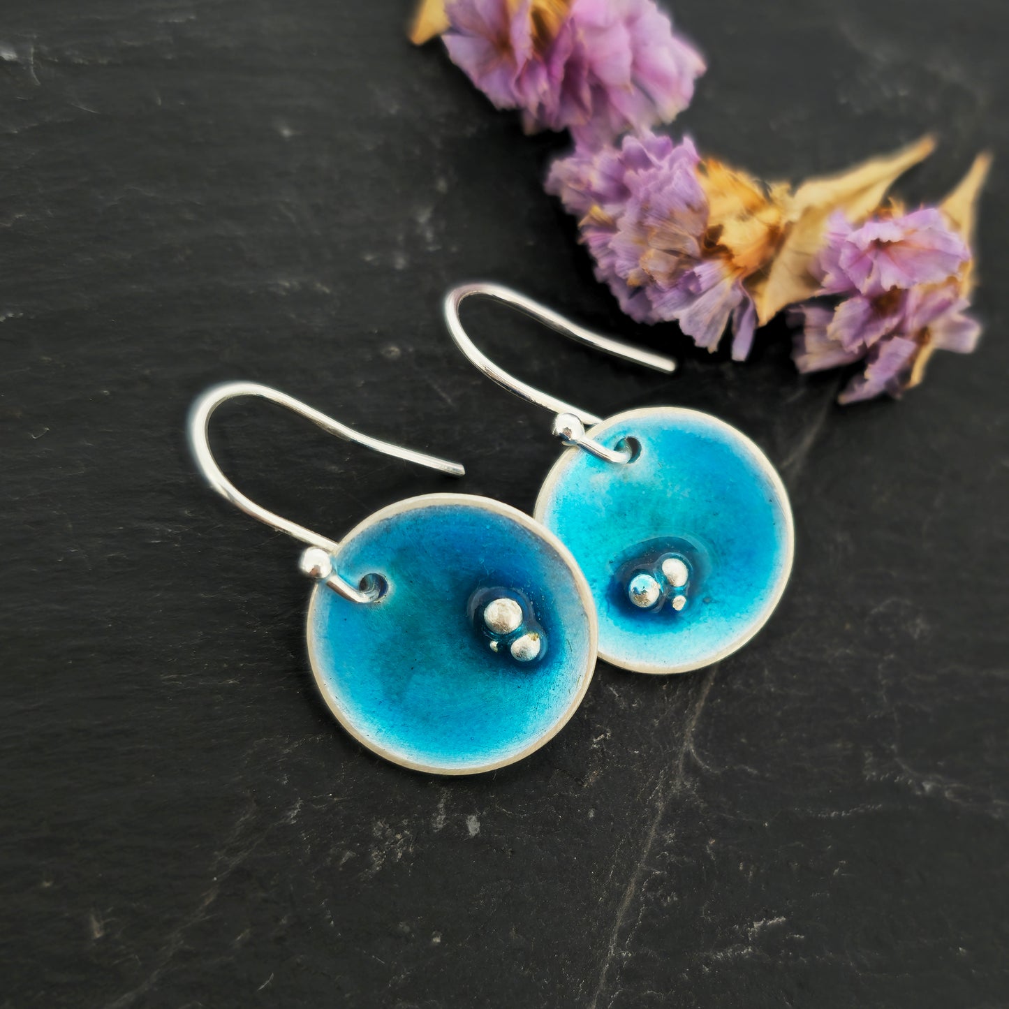 Round silver drop earrings. Each earring has 3 silver balls off-centre and a turquoise blue enamel. Suspended from silver ear hooks. Shown on slate with flowers.