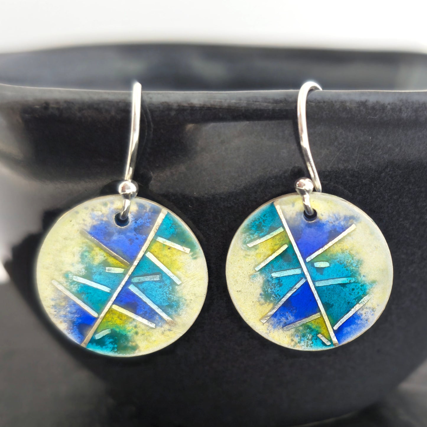 Round silver & enamel earrings with geometric design of blues, turquoise and yellow on a dark surface.