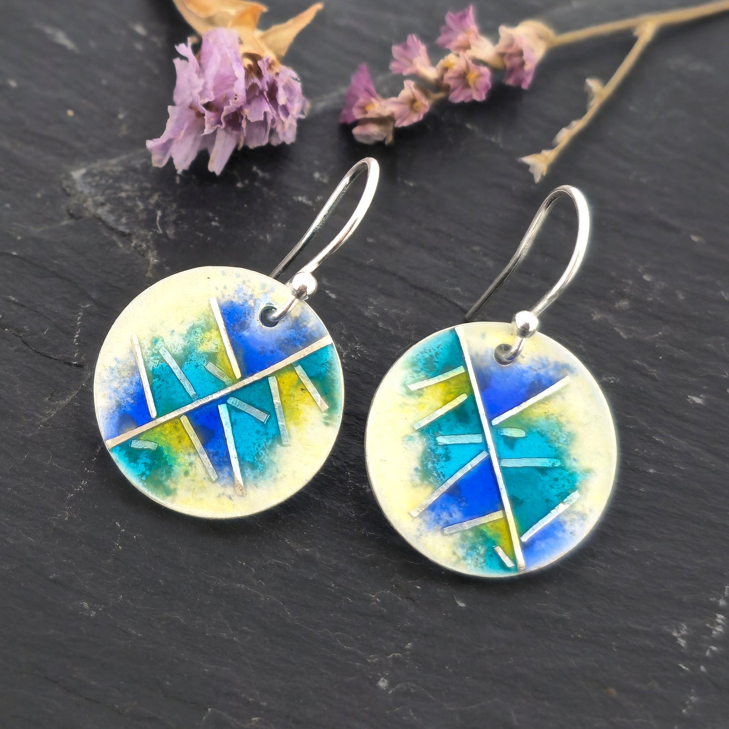 Round silver & enamel earrings with geometric design of blues, turquoise and yellow on a dark surface with dried flowers.