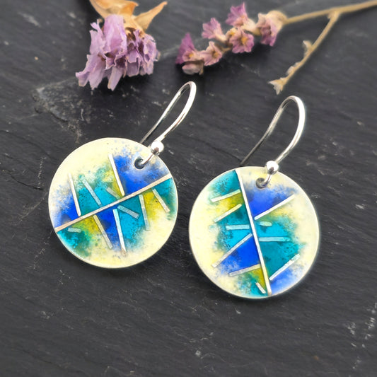 Round silver & enamel earrings with geometric design of blues, turquoise and yellow on a dark surface with dried flowers.