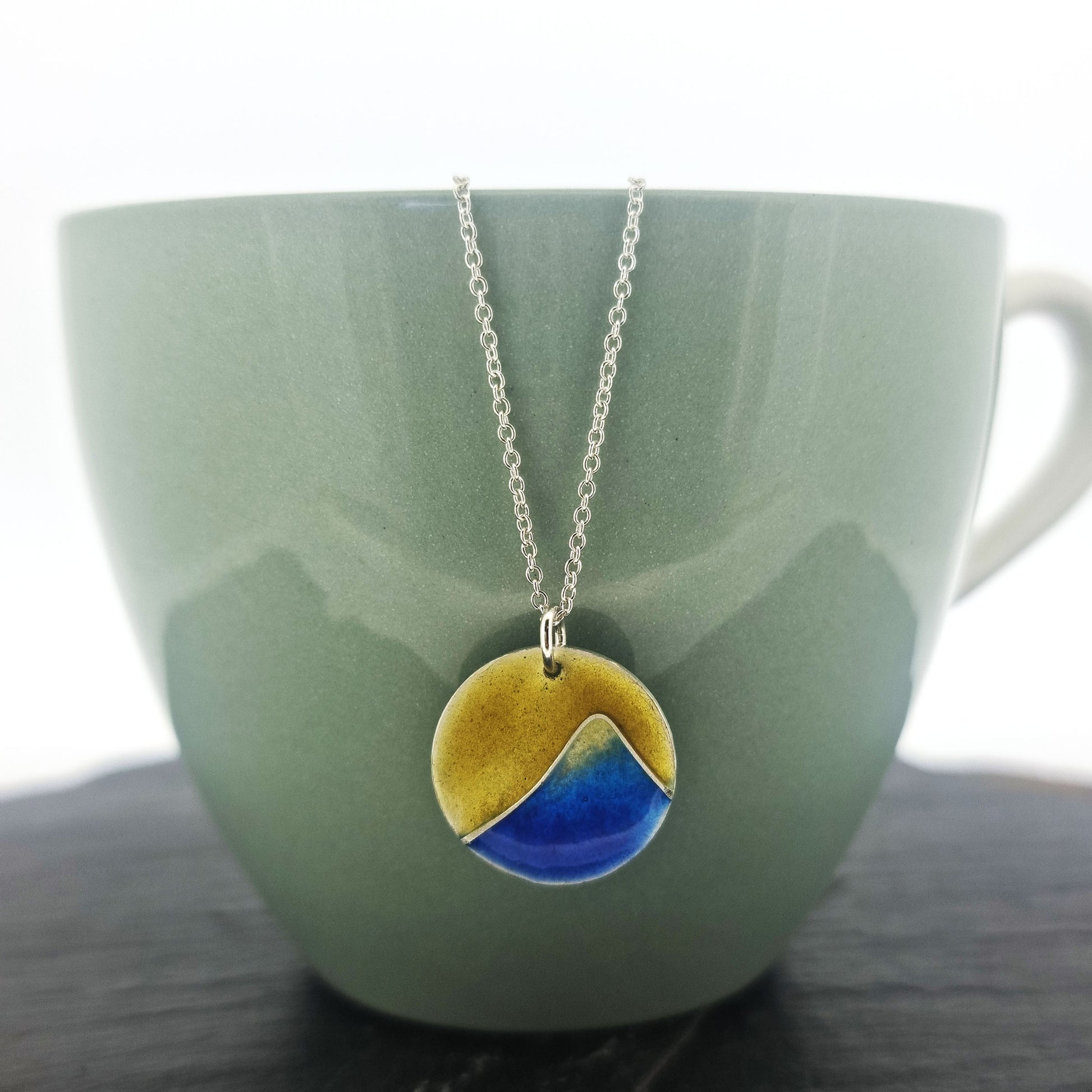 A round silver pendant with a graduated blue enamel mountain and a yellow enamel background. On a silver chain. Pictured on a cup.