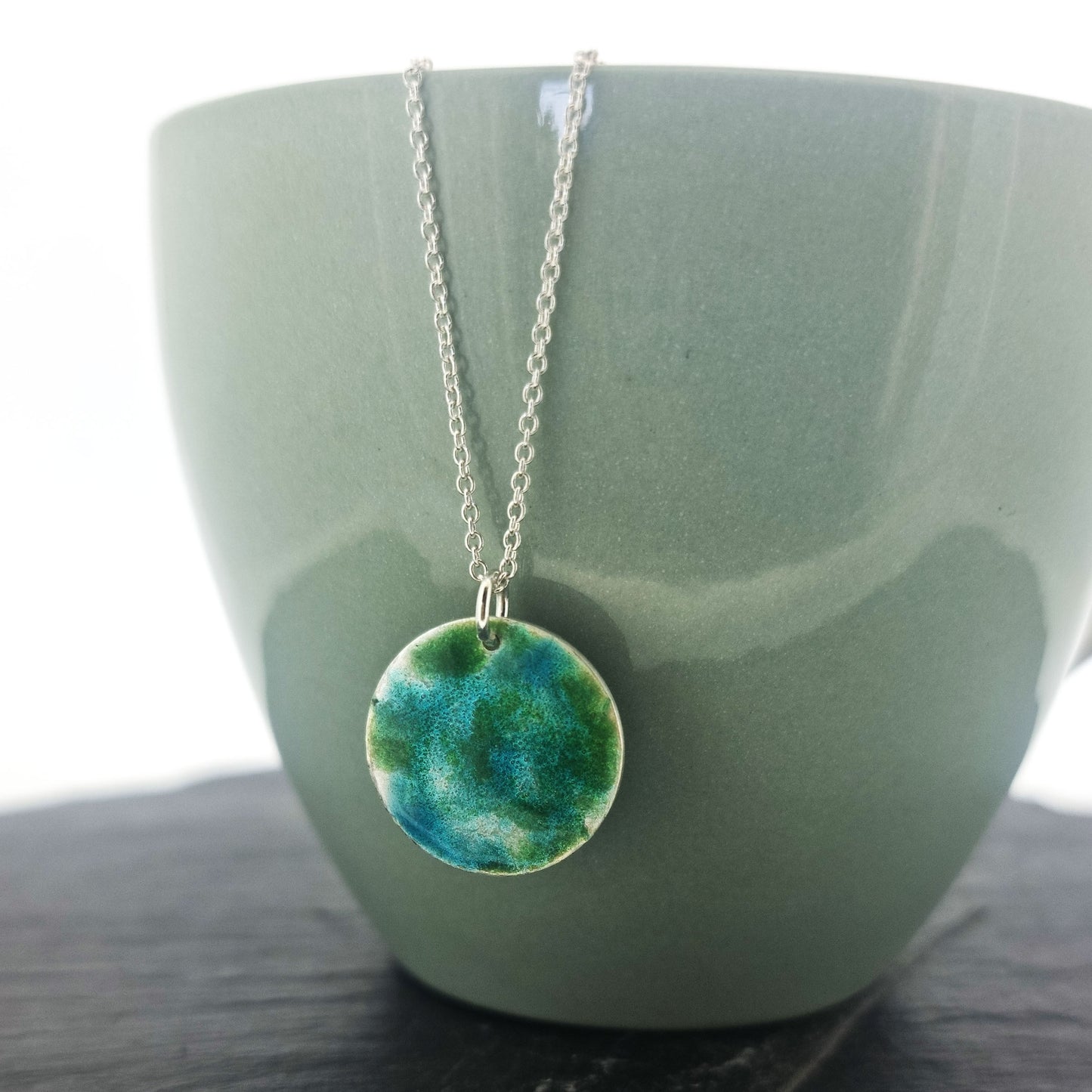 A round silver pendant with green and turquoise enamel pattern. On a silver chain. Pictured on a cup.