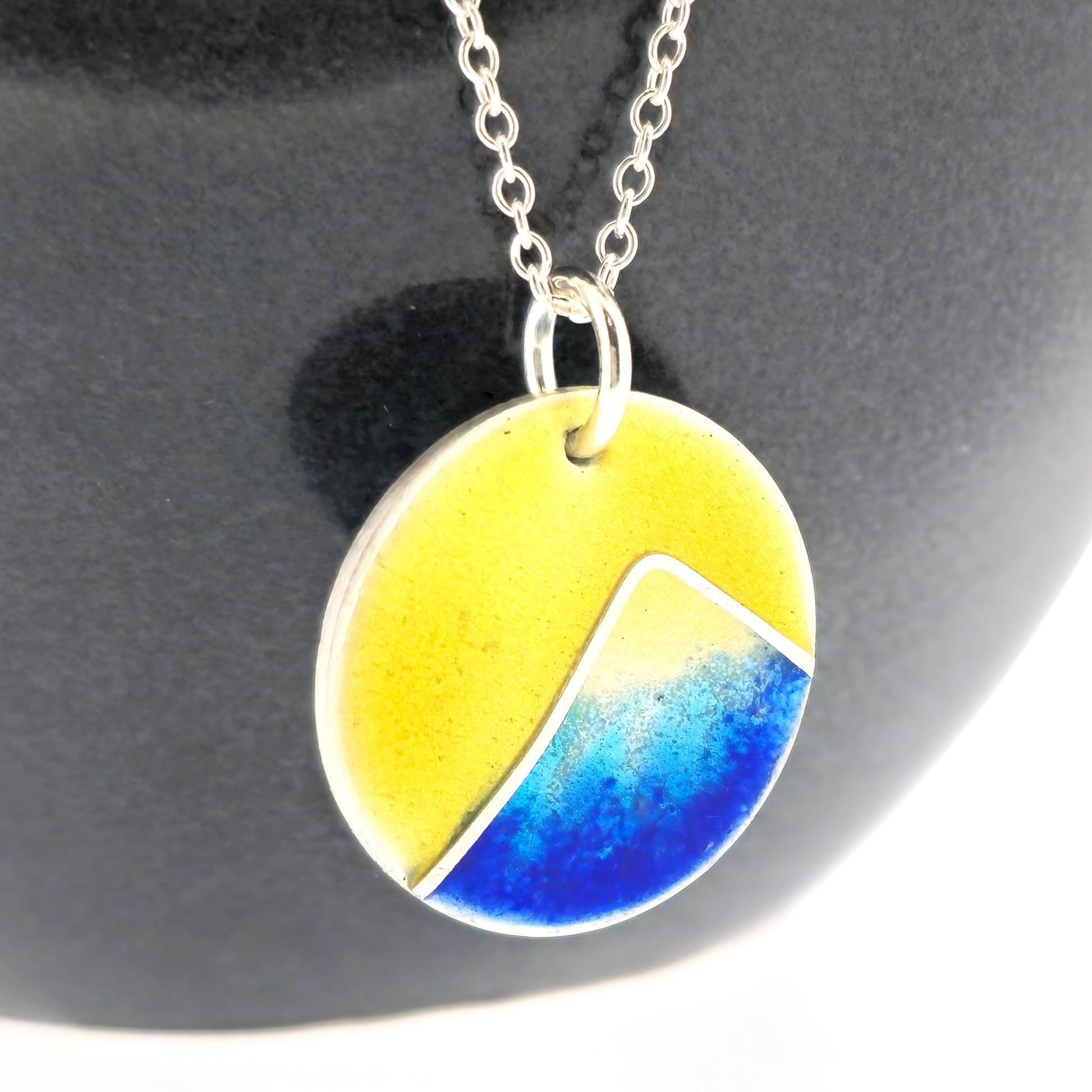 A round silver pendant with a graduated blue enamel mountain and a yellow enamel background. On a silver chain. Pictured on a black background.
