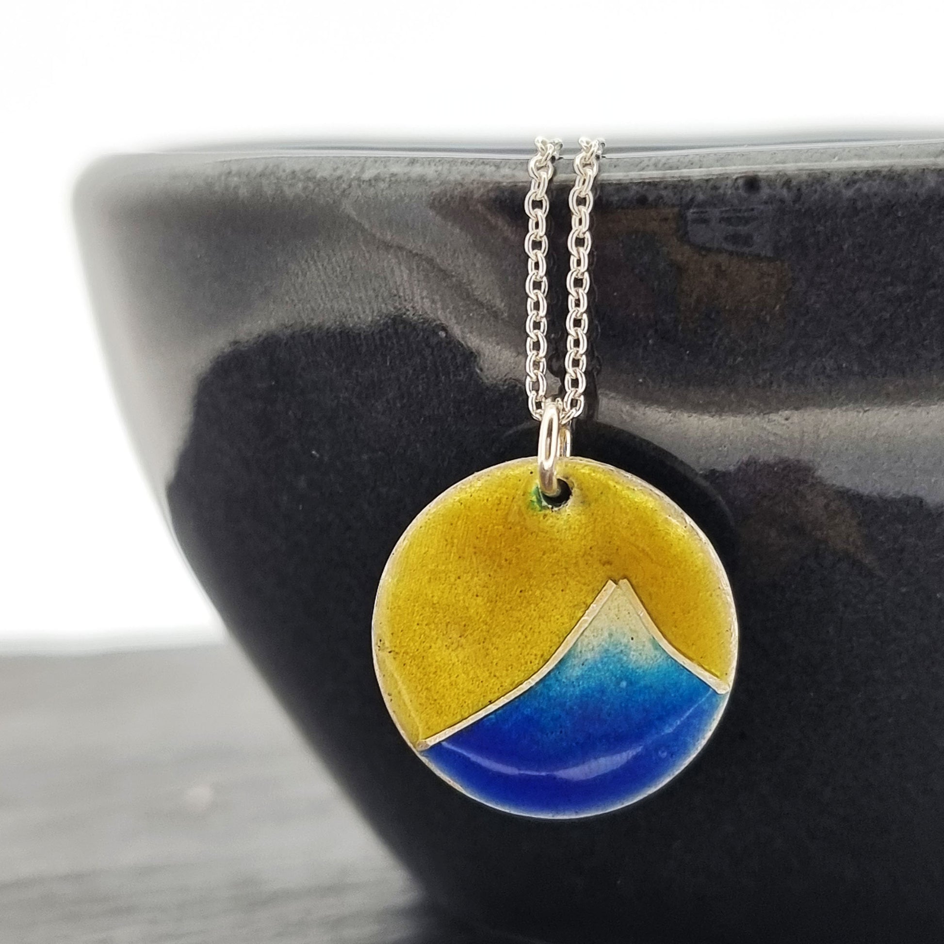 A round silver pendant with a graduated blue enamel mountain and a yellow enamel background. On a silver chain - on bowl