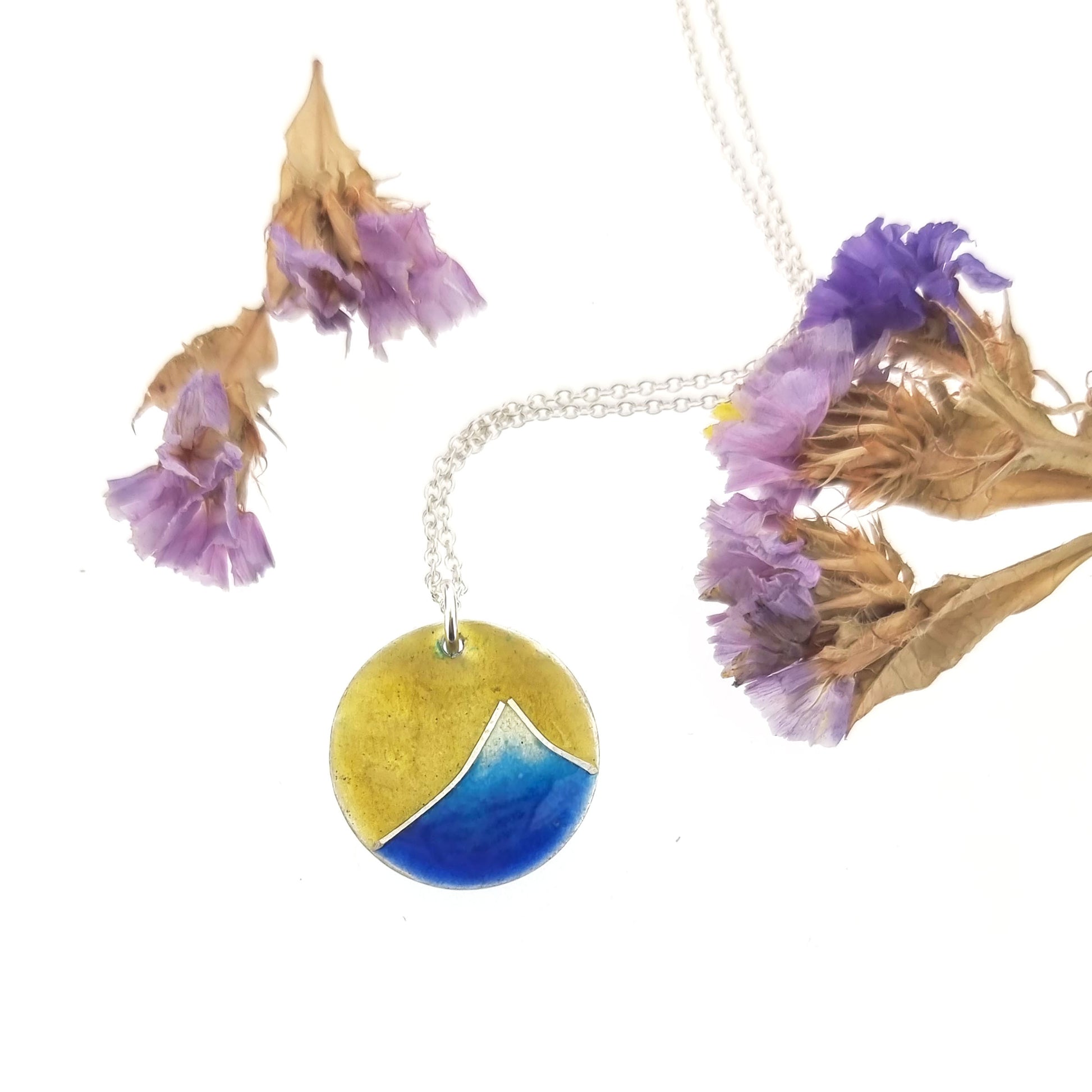 A round silver pendant with a graduated blue enamel mountain and a yellow enamel background. On a silver chain with flowers