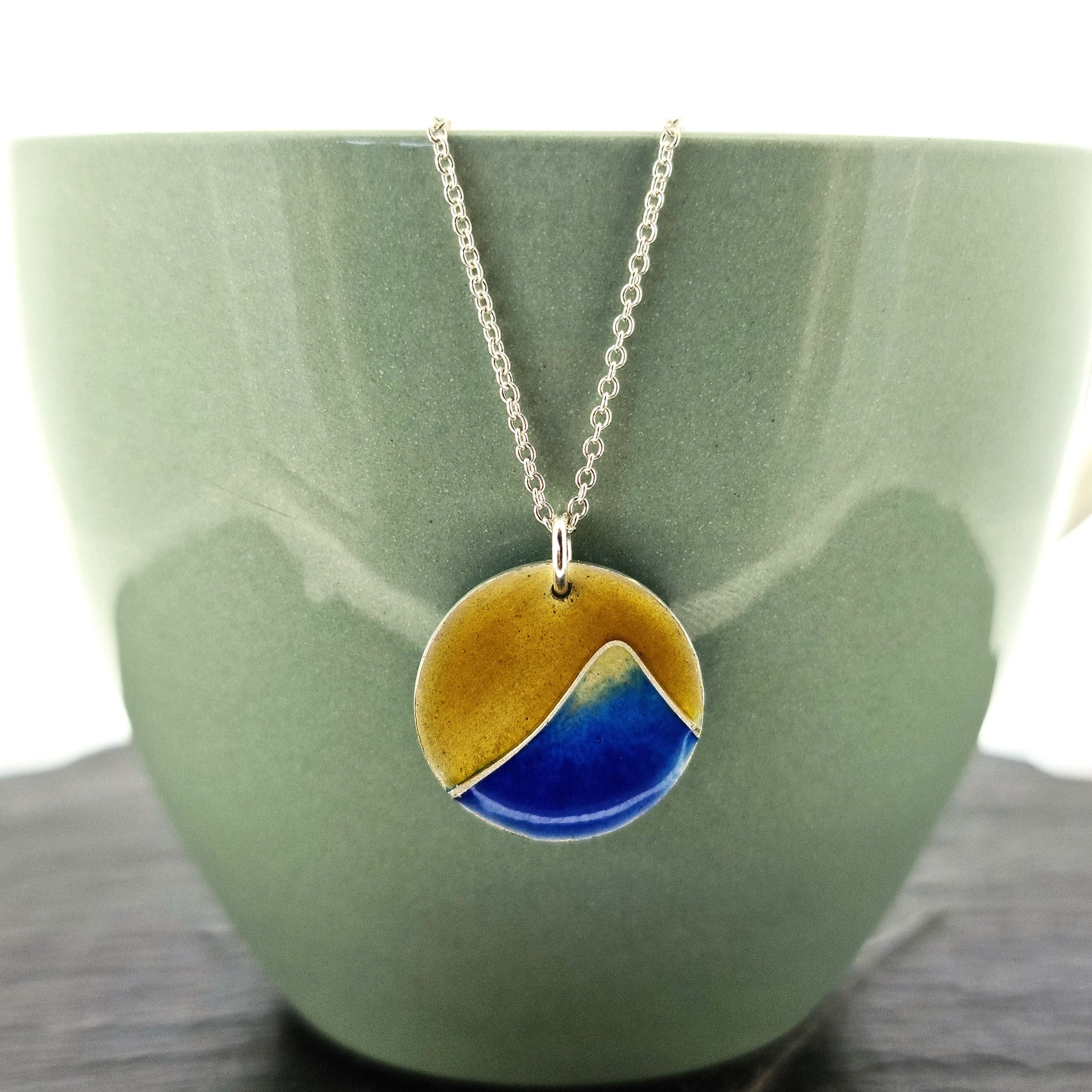 A round silver pendant with a graduated blue enamel mountain and a yellow enamel background. On a silver chain. Pictured on a cup.