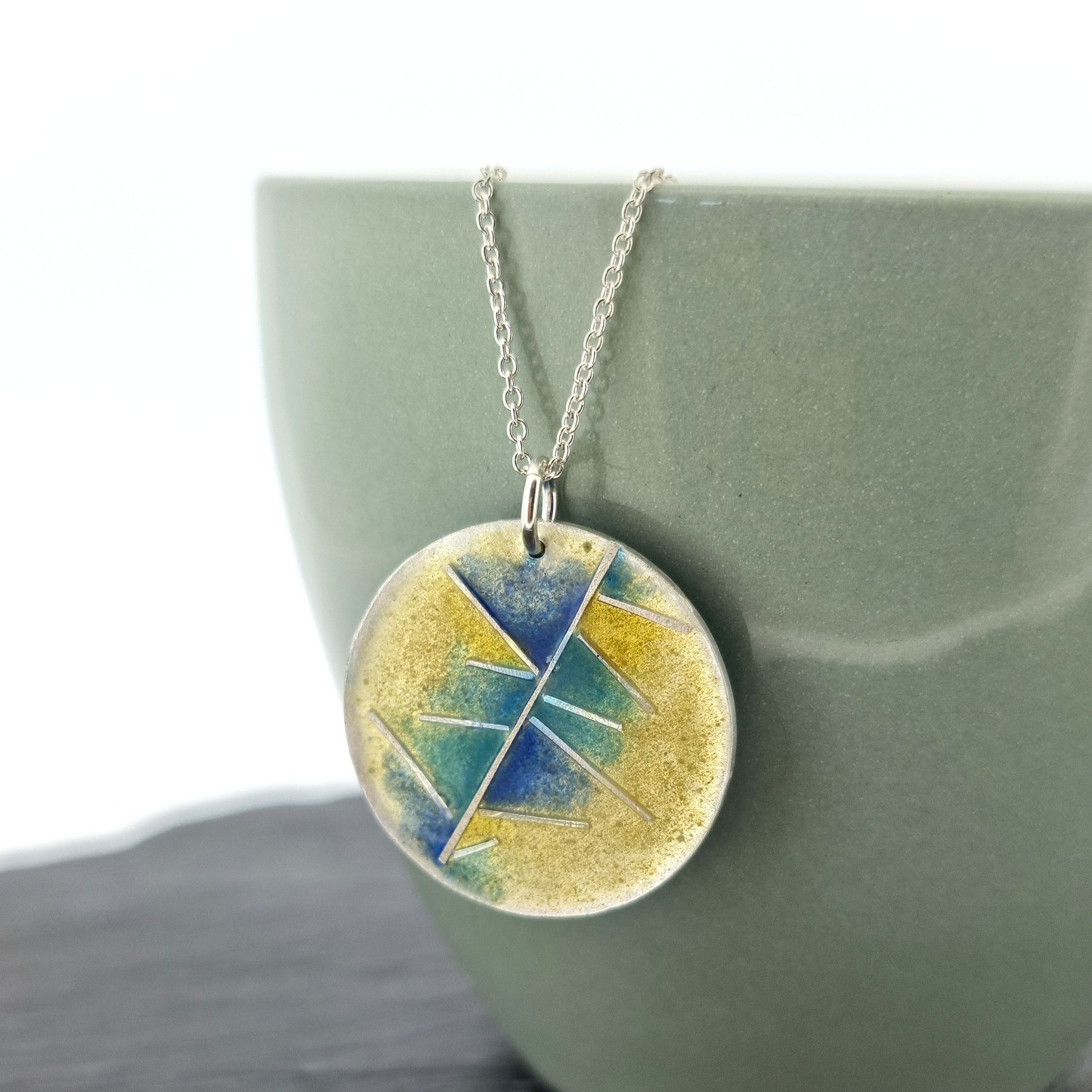 A round silver pendant with abstract silver lines and blue and turquoise enamels blending into pale yellow. Suspended from a silver chain. Pictured on a cup.