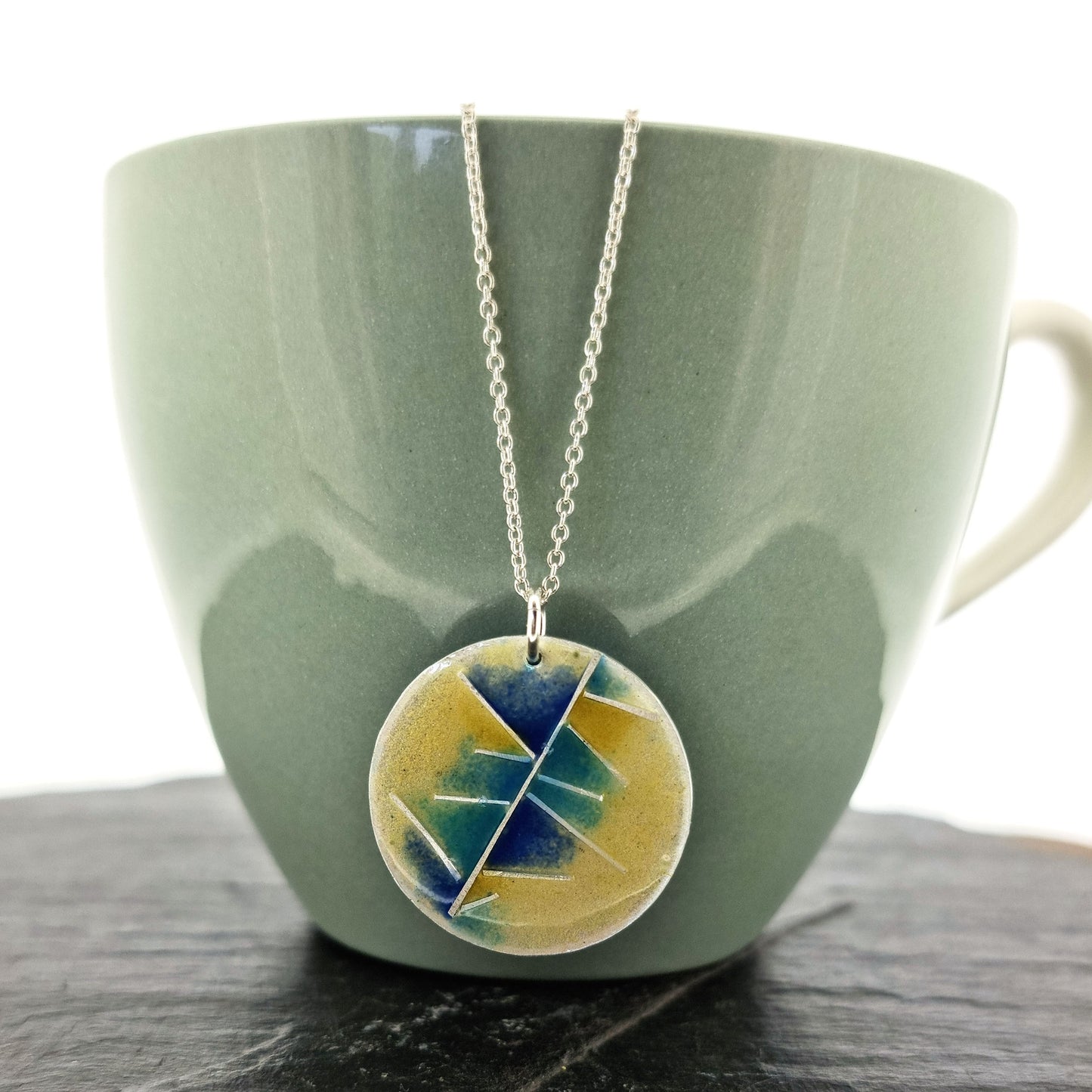 A round silver pendant with abstract silver lines and blue and turquoise enamels blending into pale yellow. Suspended from a silver chain. Pictured on a cup.