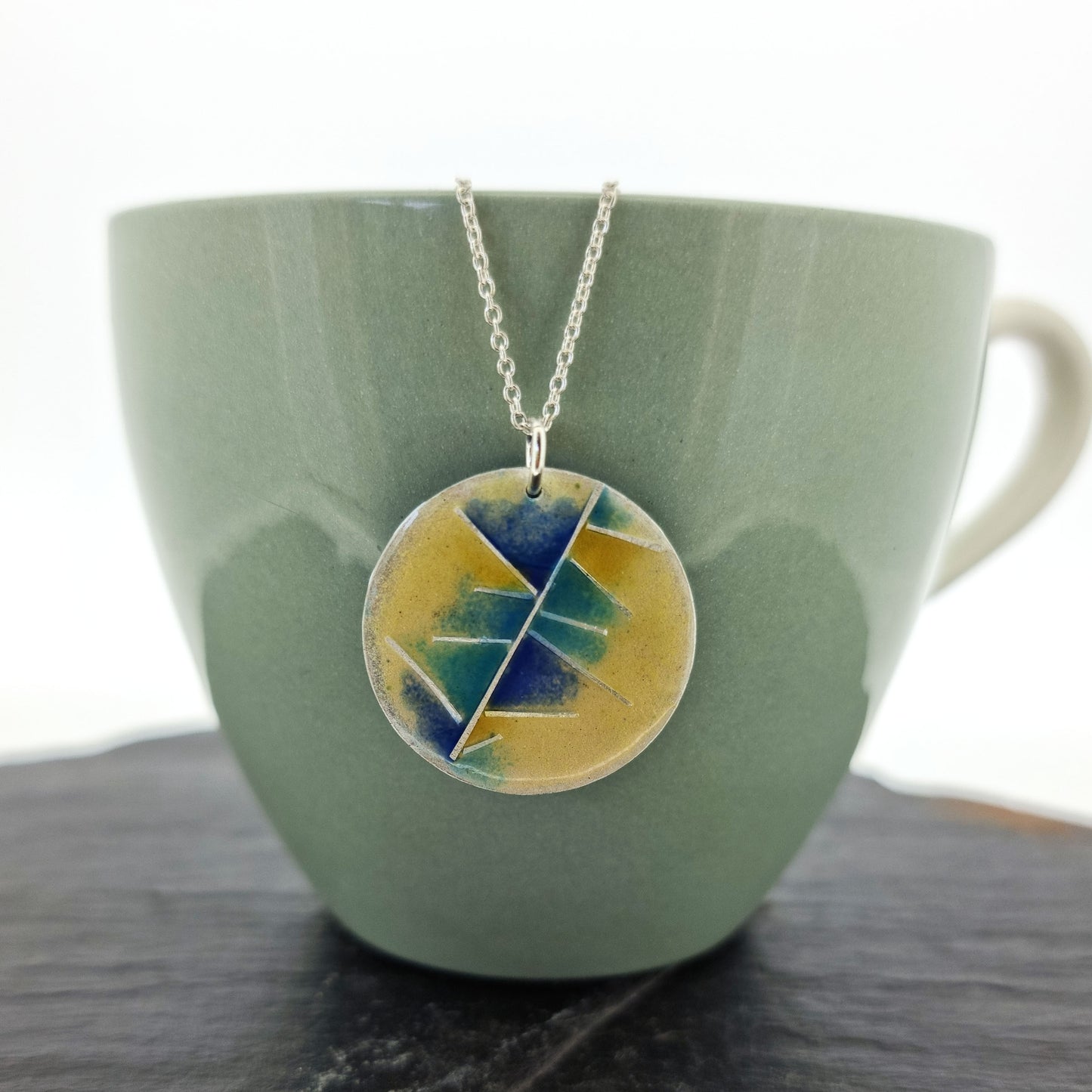 A round silver pendant with abstract silver lines and blue and turquoise enamels blending into pale yellow. Suspended from a silver chain. Pictured on a cup.