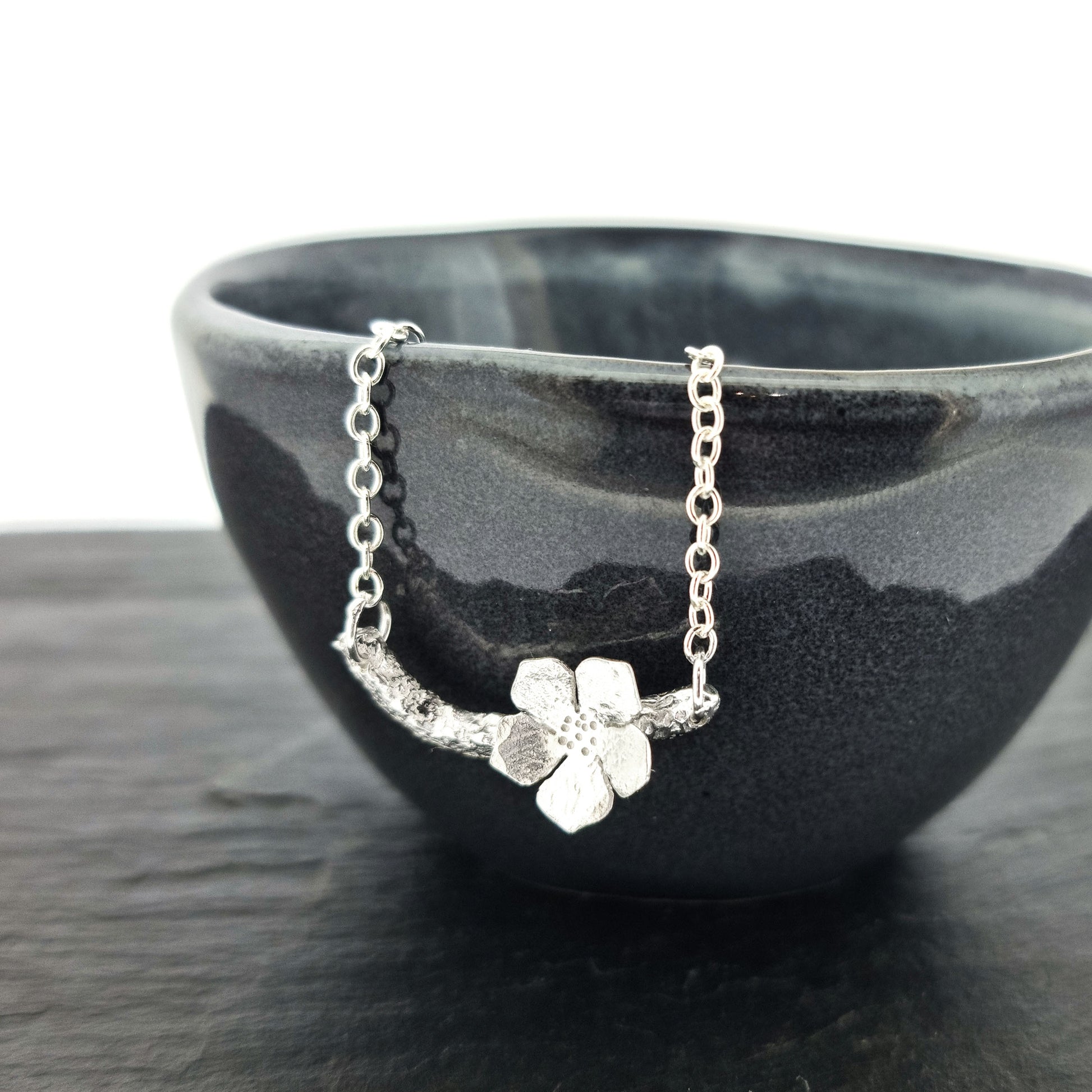 Silver necklace with small branch and a single 5 petal flower on it. Shown on a black bowl.