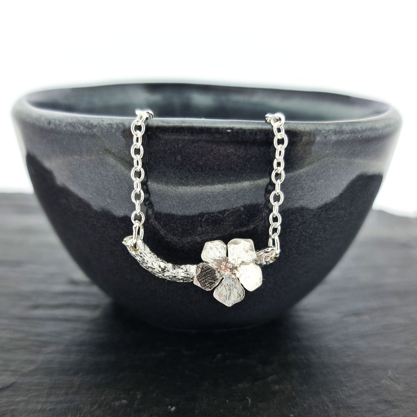 Silver necklace with small branch and a single 5 petal flower on it. Shown on a black bowl.