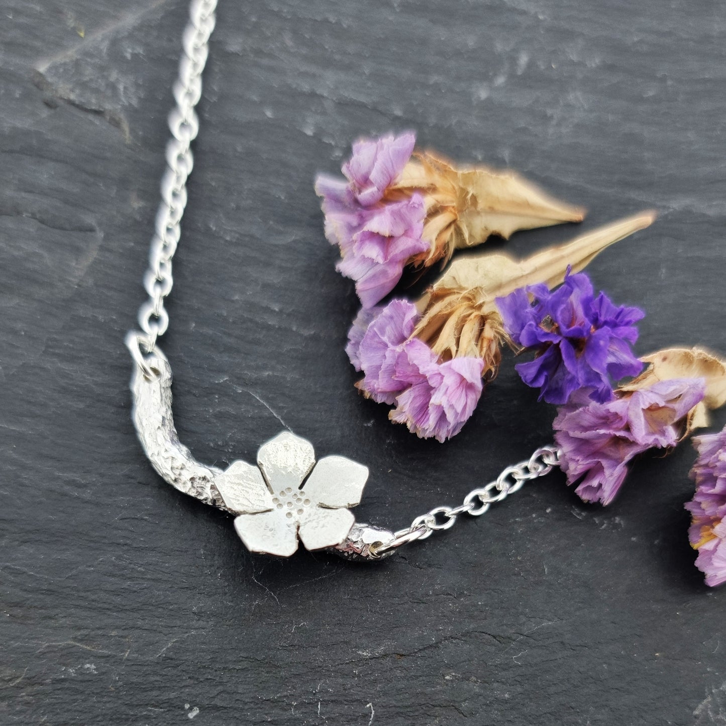 Silver necklace with small branch and a single 5 petal flower on it. Pictured with flowers. 