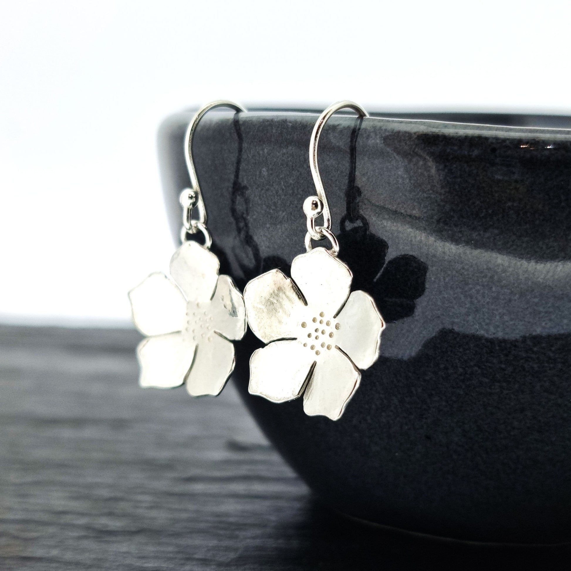 Silver five petal flower drop earrings. Pictured on a black bowl.