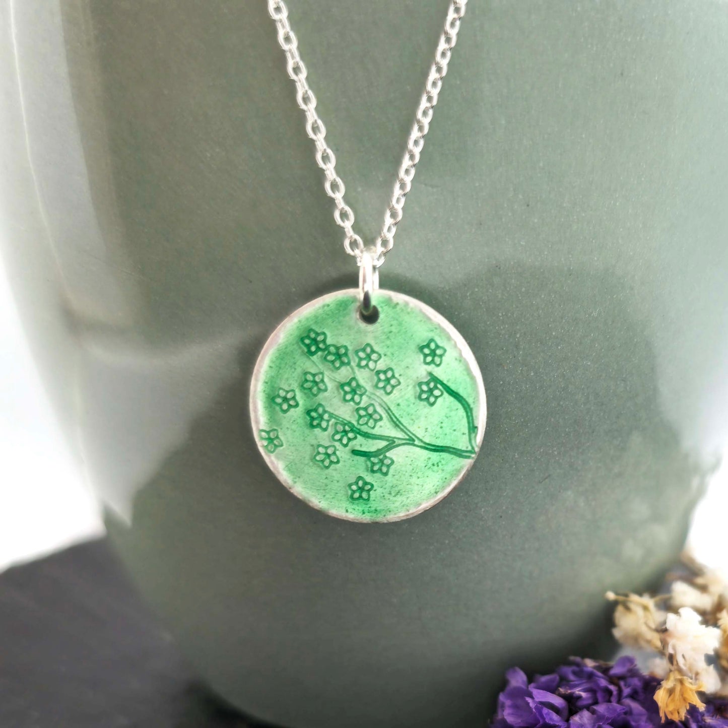 Silver necklace with a round green enamel pendant featuring floral designs on a green surface with flowers in the background.