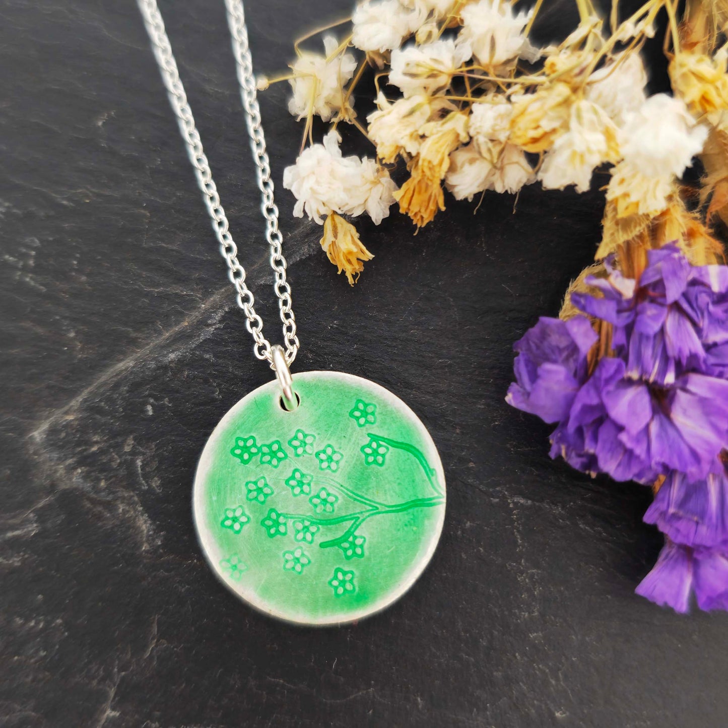 Silver necklace with a round green enamel pendant featuring floral designs on a black surface with flowers.