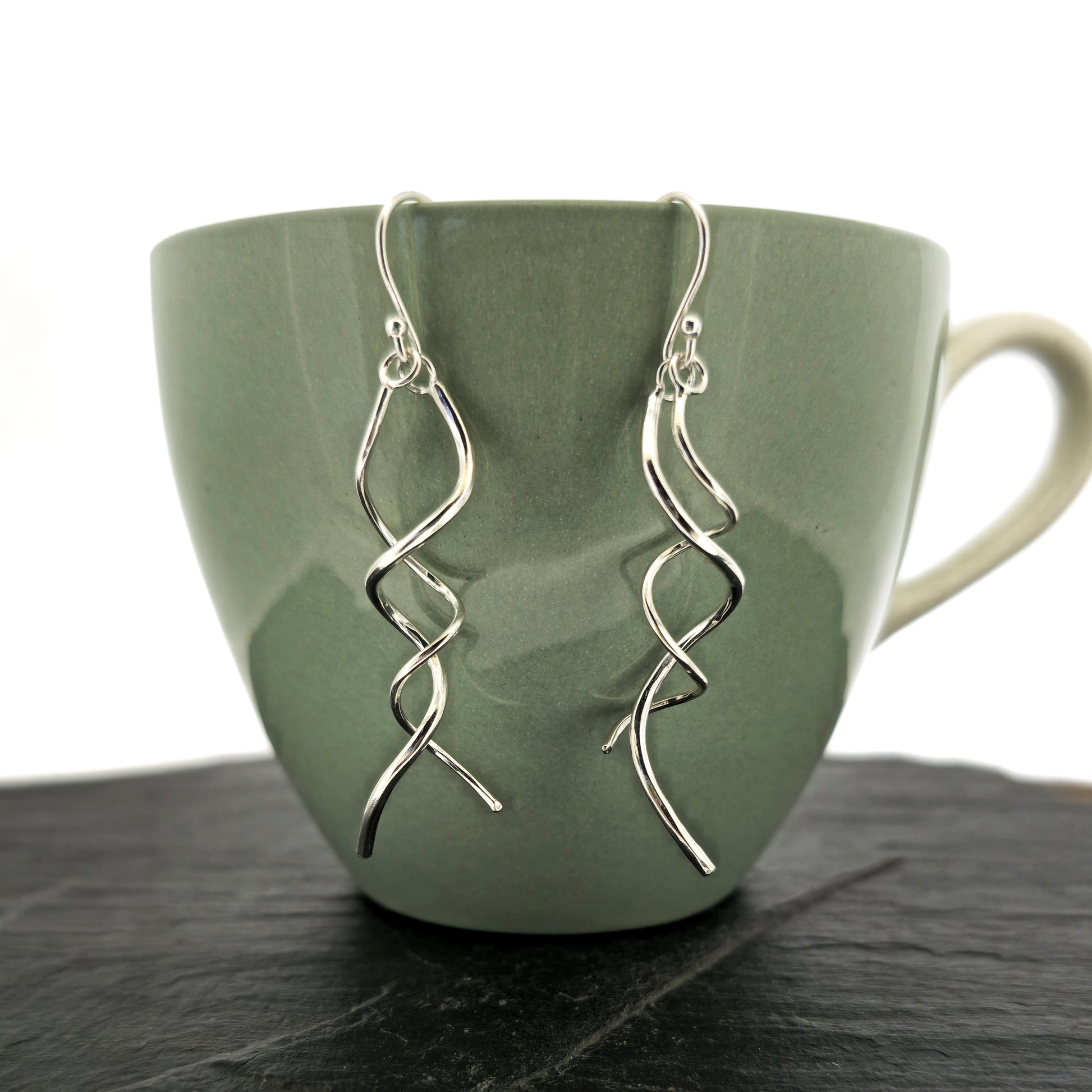 Silver spiral drop earrings featuring 2 curled strands intertwined. Shown on a cup.