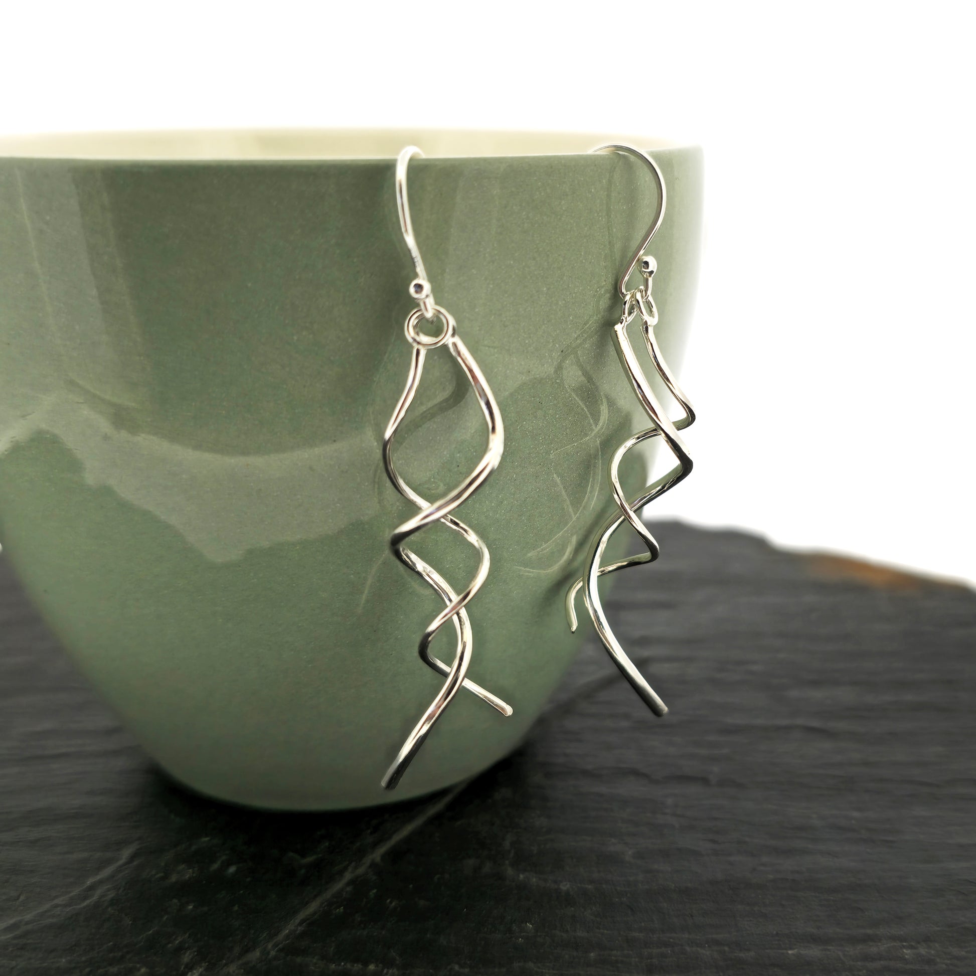 Silver spiral drop earrings featuring 2 curled strands intertwined. Shown on a cup.