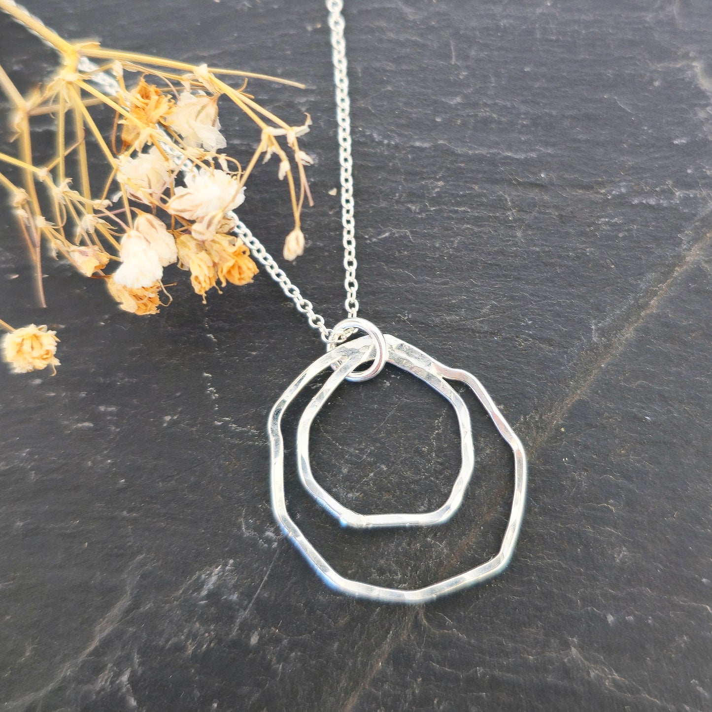 A handmade silver pendant consisting of two organic rustic style open circles of different sizes suspended from a silver chain. On a slate background with dried flowers. Mijoux Jewellery.
