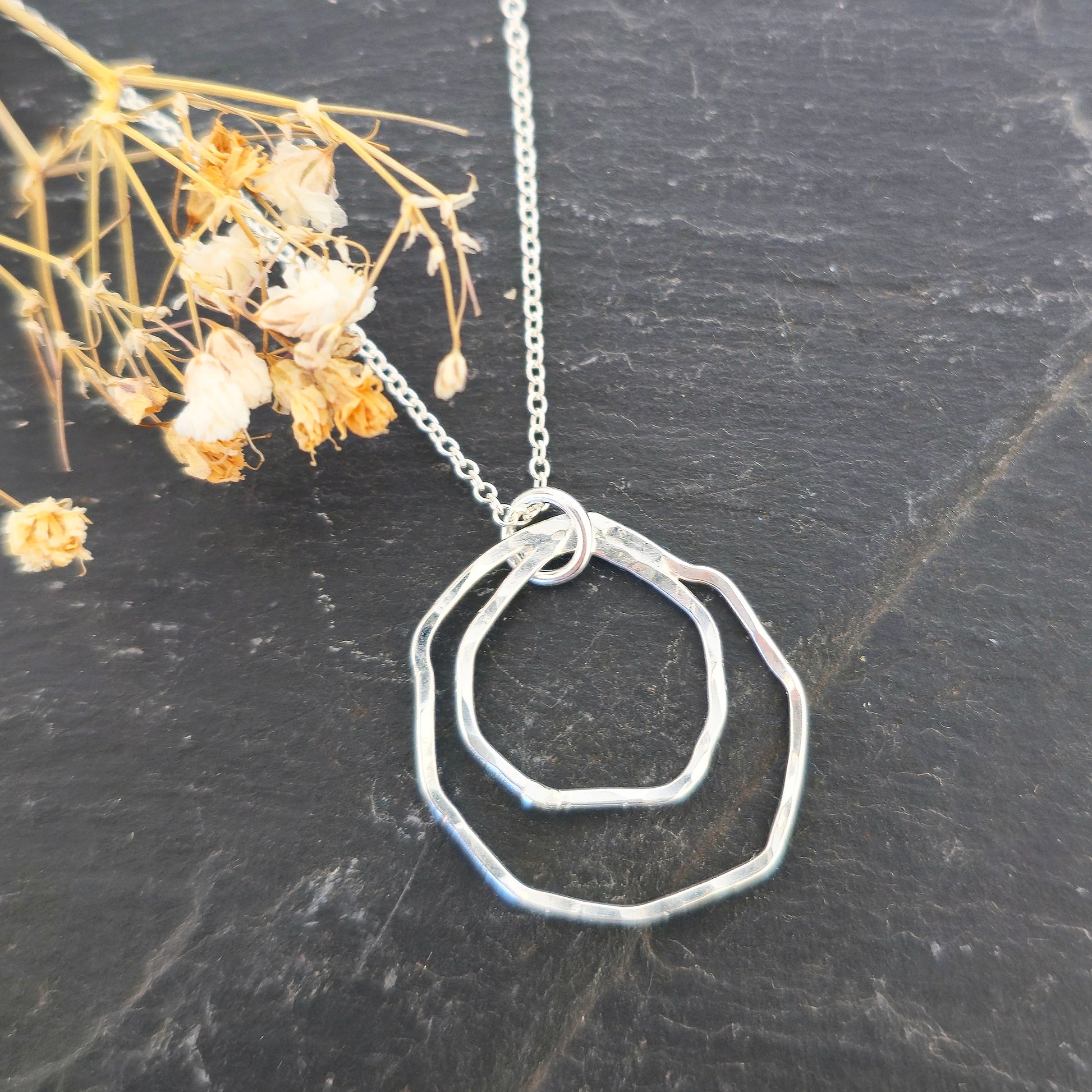 A handmade silver pendant consisting of two organic rustic style open circles of different sizes suspended from a silver chain. On a slate background with dried flowers. Mijoux Jewellery.
