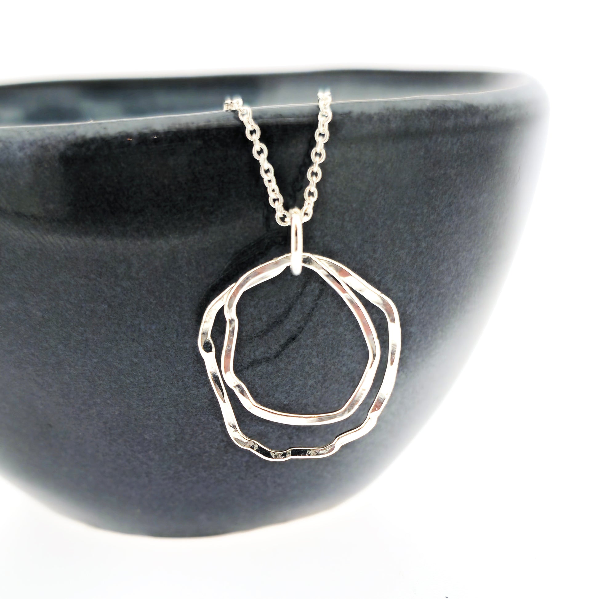 A silver pendant consisting of two organic rustic style open circles of different sizes suspended from a silver chain. Shown on a black bowl.