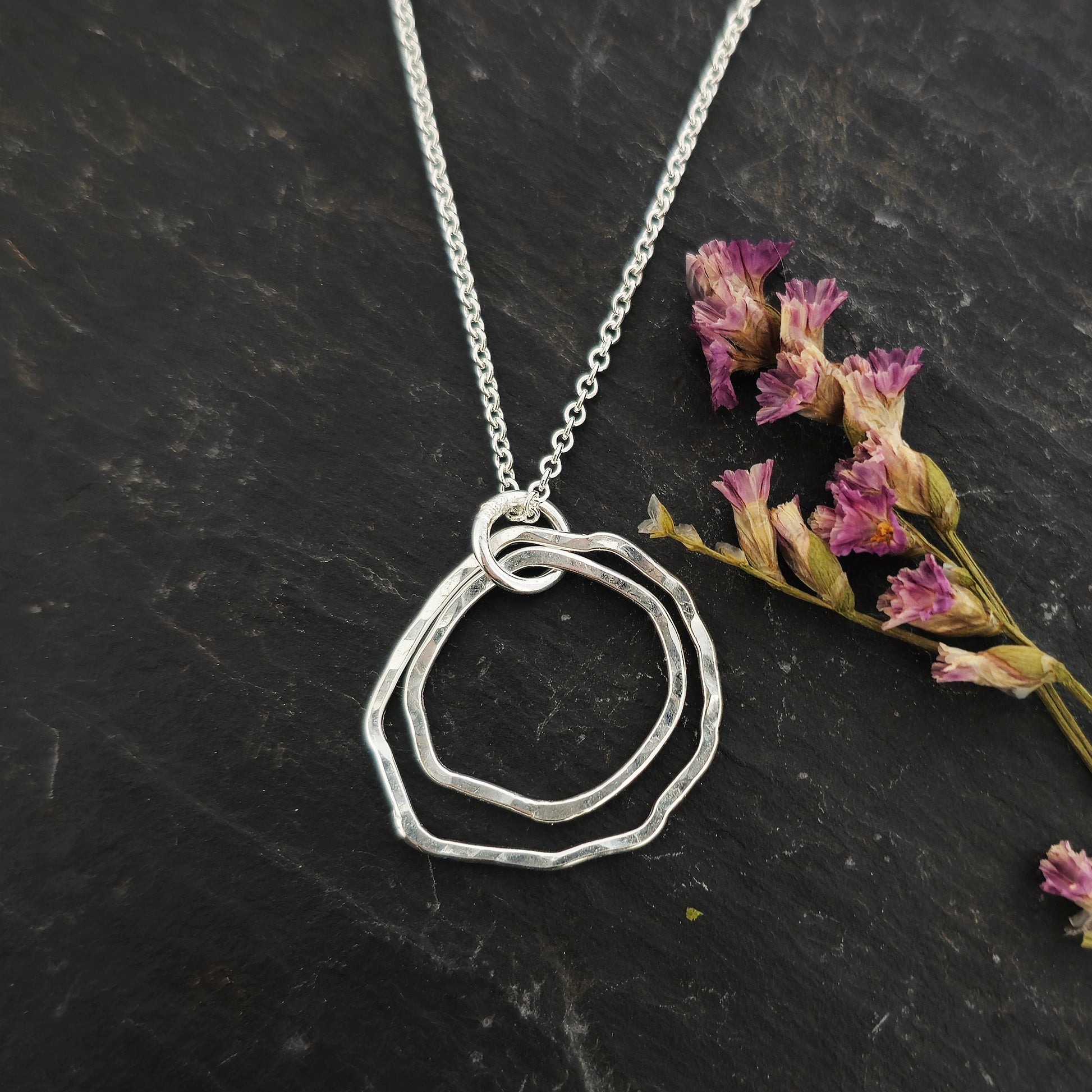 A silver pendant consisting of two organic rustic style open circles of different sizes suspended from a silver chain. Shown on slate with flowers.