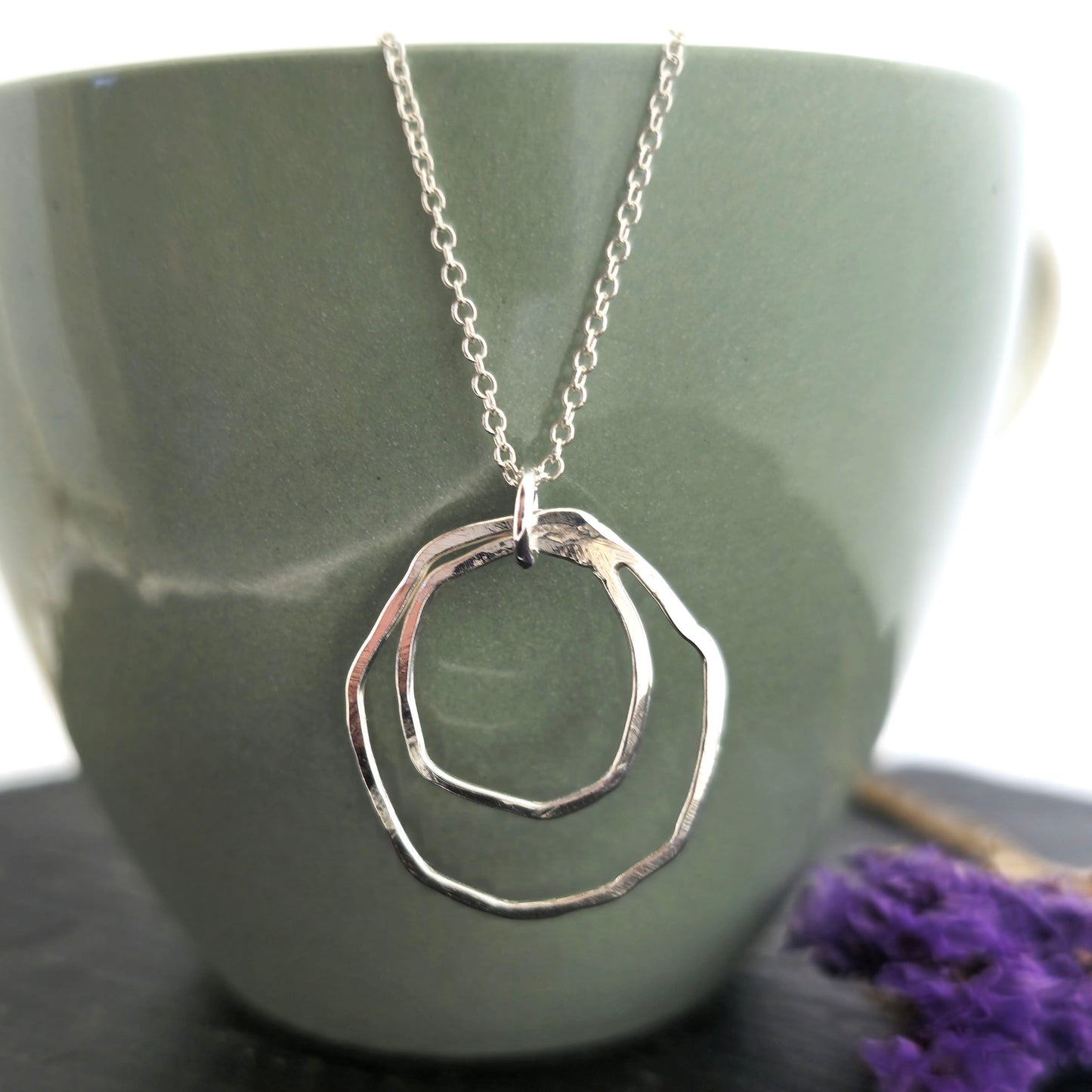 A handmade silver pendant consisting of two organic rustic style open circles of different sizes suspended from a silver chain. On a green cup with dried flowers. Mijoux Jewellery.