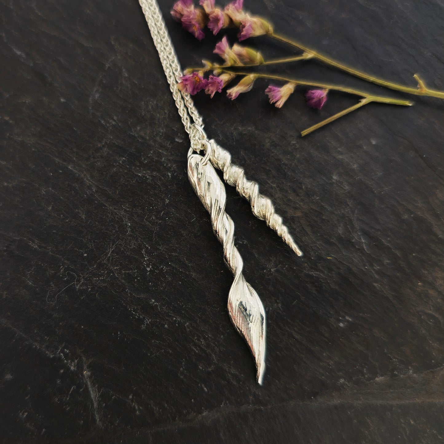 Two silver twist pendants of different sizes suspended from a long silver chain. Shown on slate with flowers.