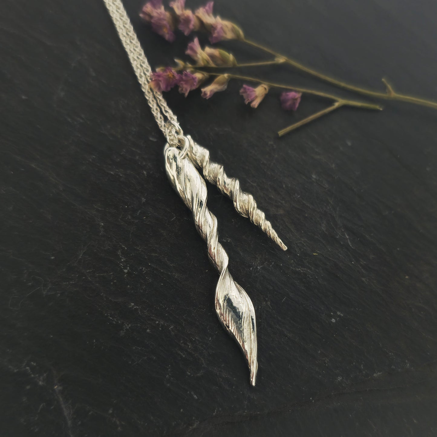 Two silver twist pendants of different sizes suspended from a long silver chain. Shown on slate with flowers.