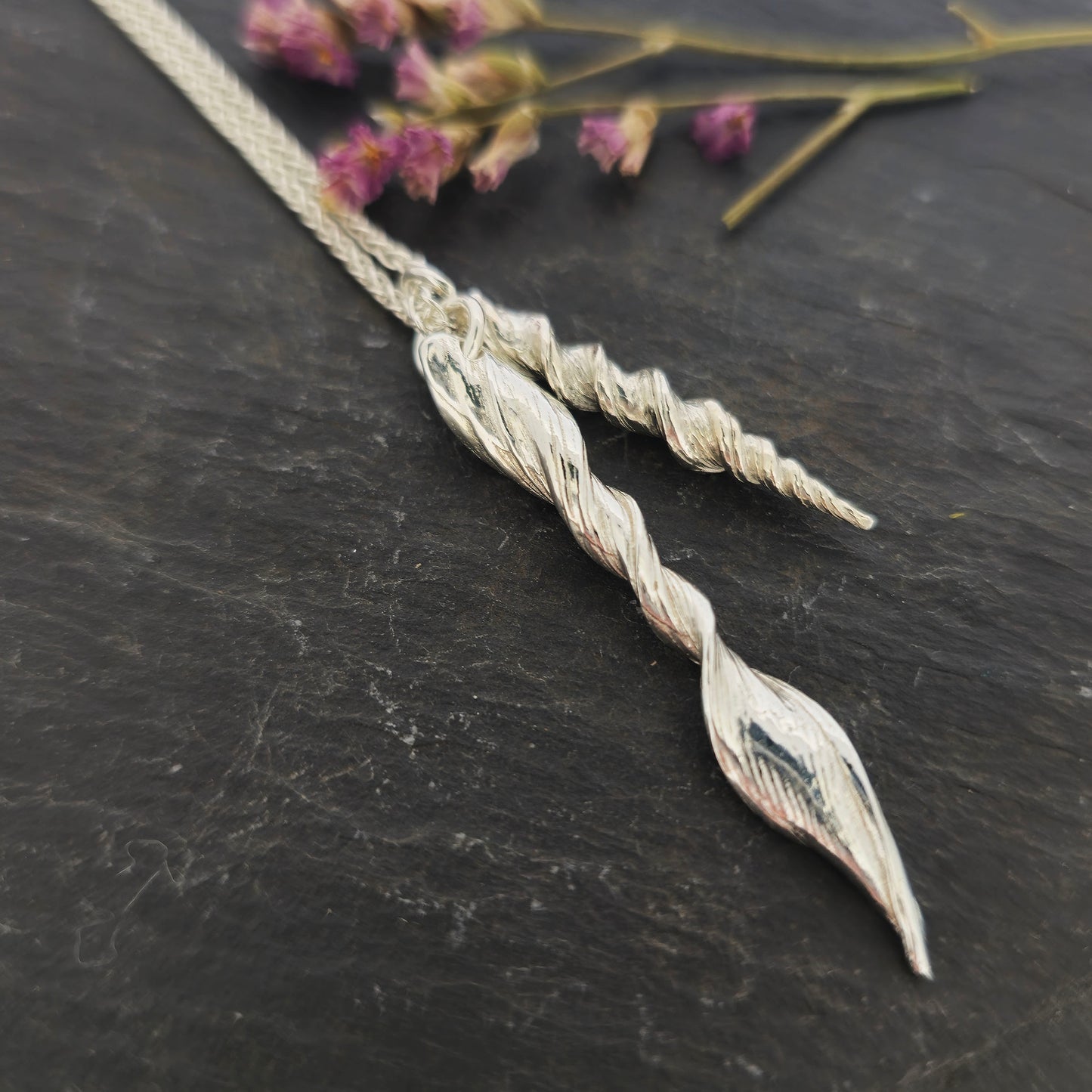 Two silver twist pendants of different sizes suspended from a long silver chain. Shown on slate with flowers.