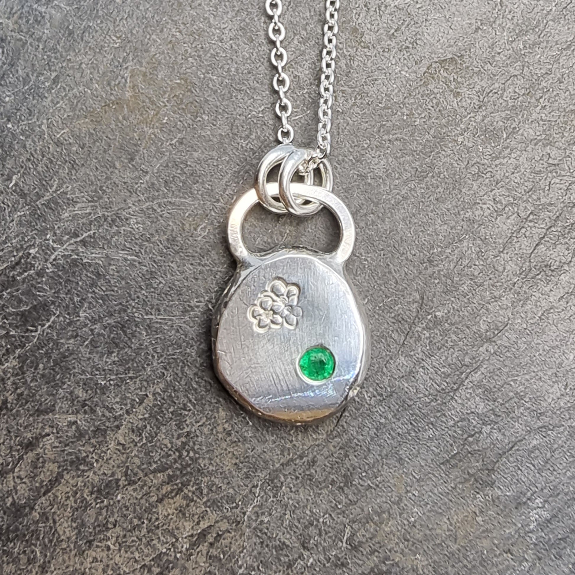 Round silver pendant necklace with a hawthorn flower engraved on it and a flush set green emerald gemstone. On a silver chain.