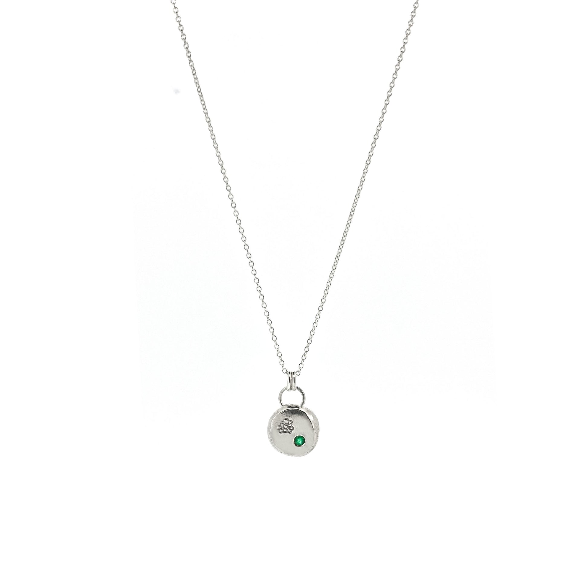 Round silver pendant necklace with a hawthorn flower engraved on it and a flush set green emerald gemstone. On a silver chain.